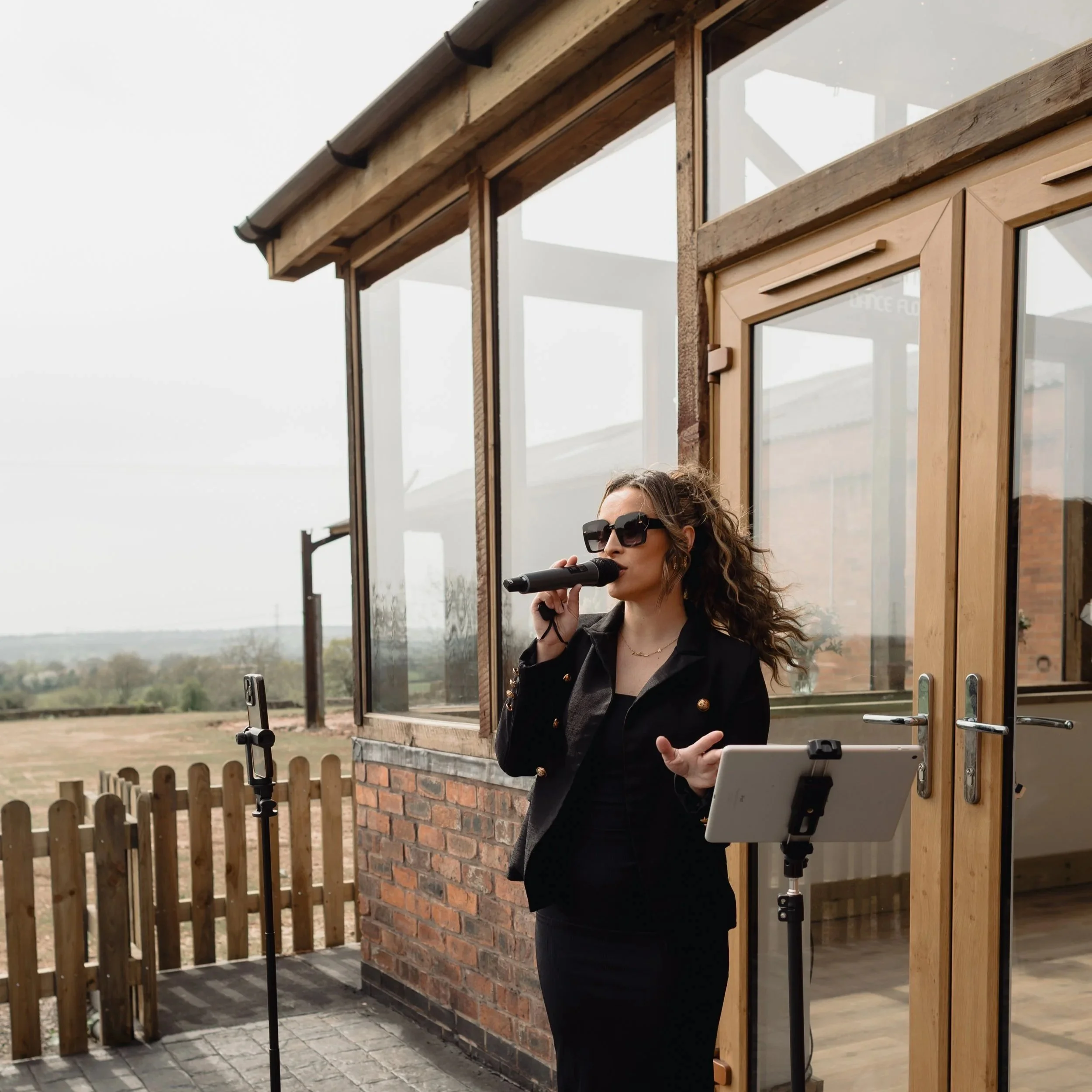A woman wearing large sunglasses and a black blazer stands outside near a wooden house with large glass windows and a wooden fence. She appears to be speaking or singing into a microphone on a stand.