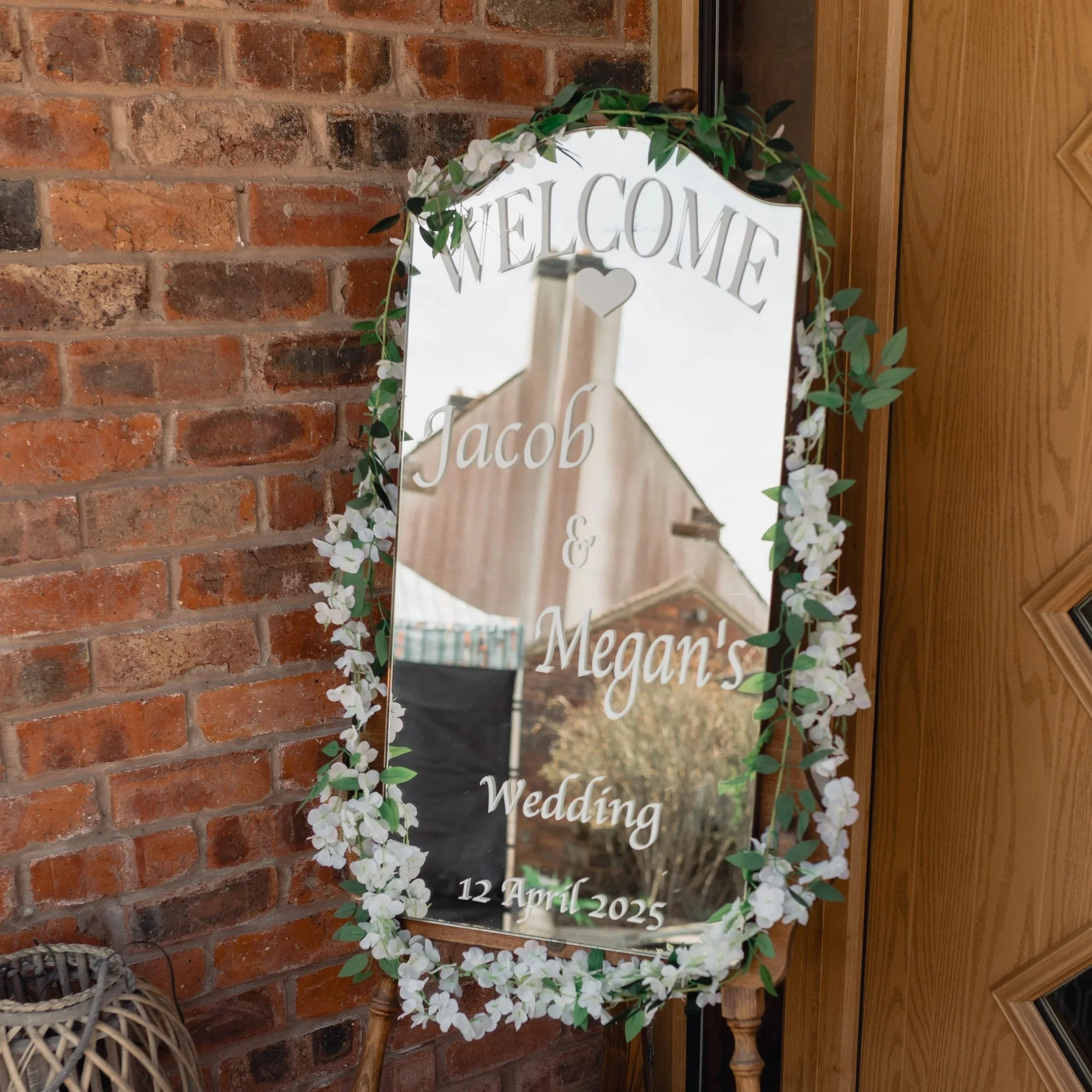 A mirror decorated with white flowers and green leaves, with the words 'Welcome', 'Jack', and 'Megan' visible, along with a wedding date of April 12, 2025. The mirror is mounted on a brick wall next to a wooden surface.
