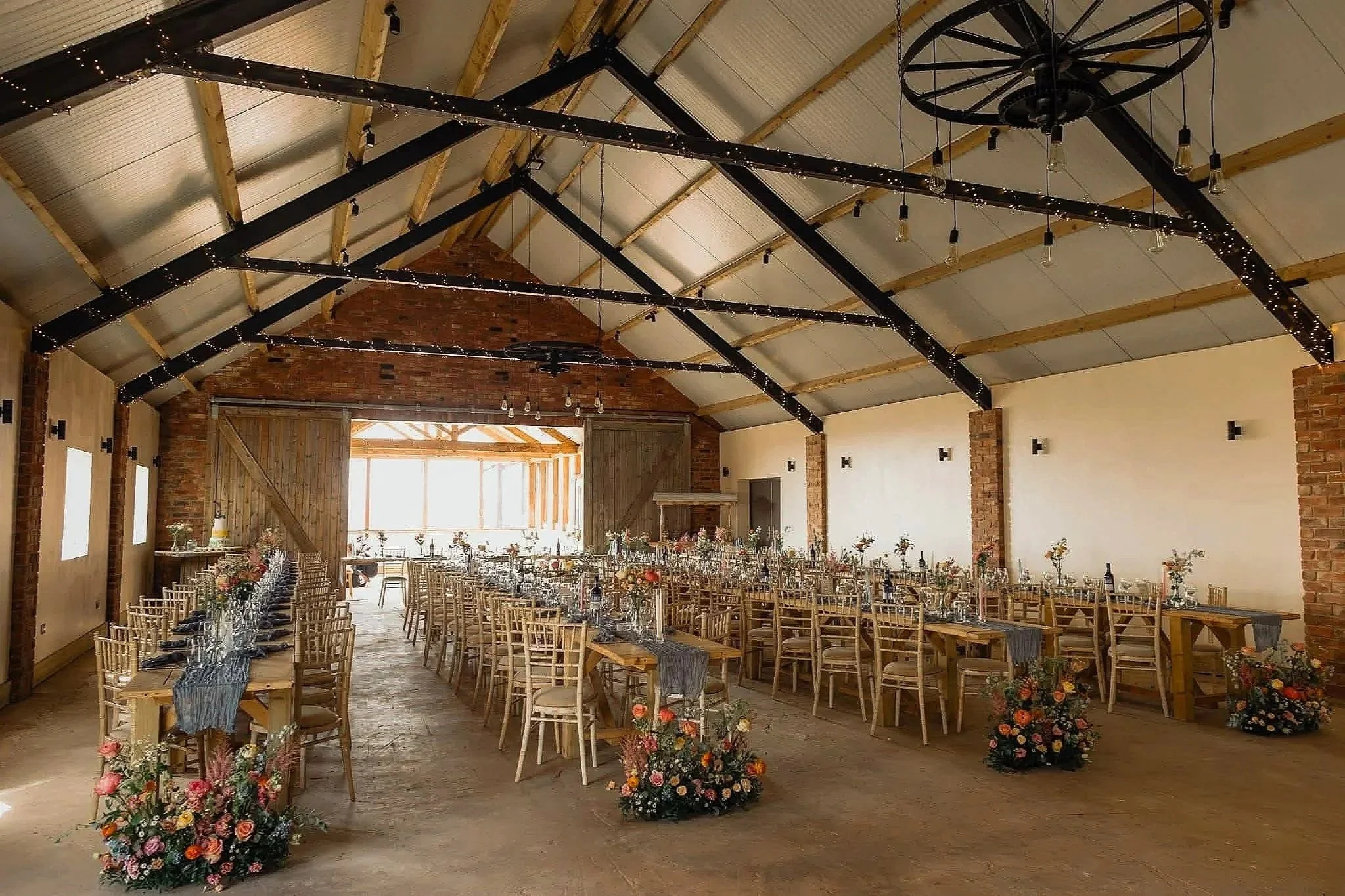 Large rustic barn interior decorated for a wedding reception with long tables, wooden chairs, floral centerpieces, and string lights hanging from the ceiling beams.