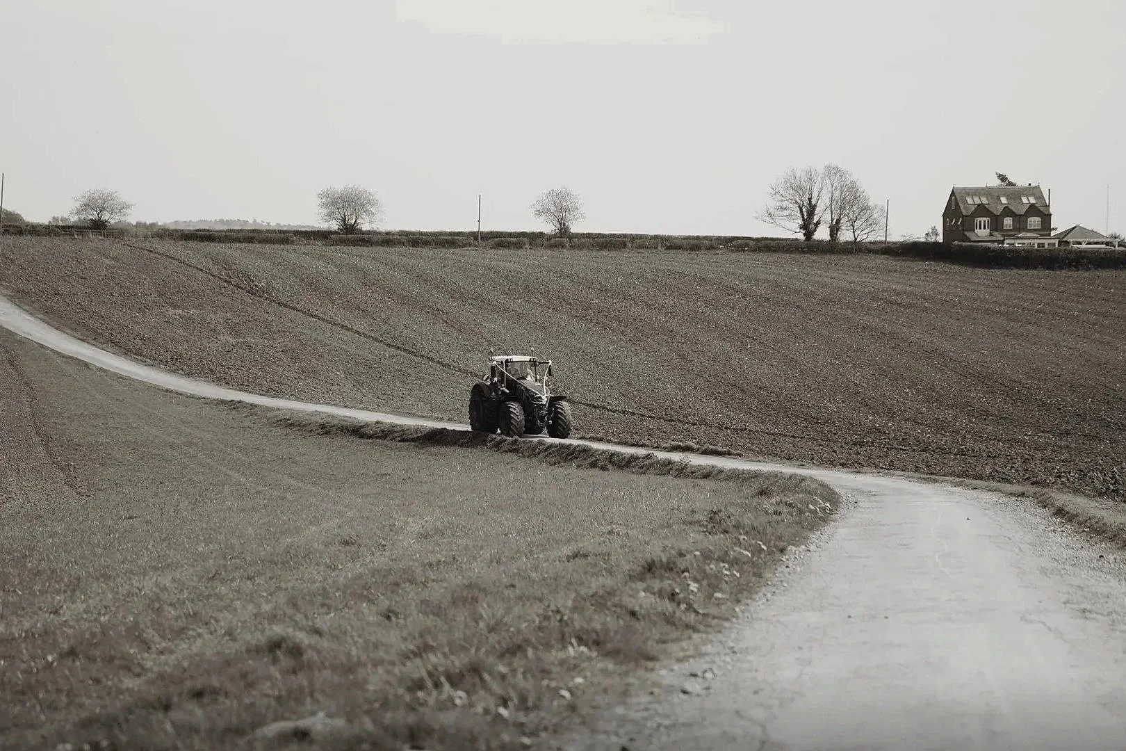 A tractor plowing a field on a rural farm with a house and trees in the background.