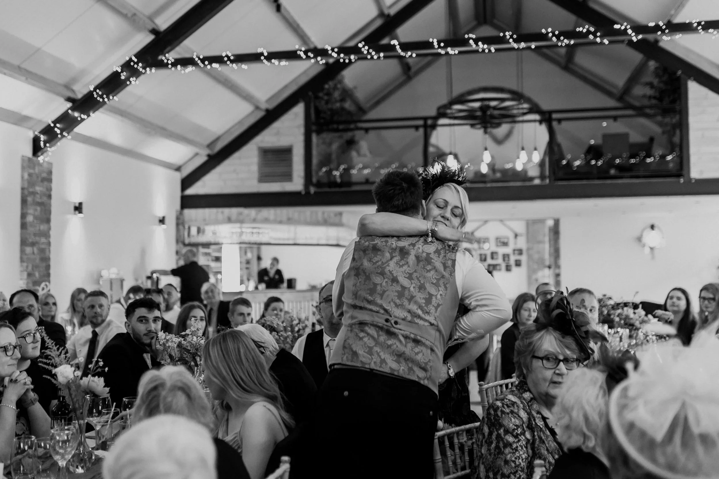 A black-and-white photo of a wedding reception with guests seated at ta bles decorated with flowers. A couple is dancing in the center, embracing each other, in a warmly lit venue with string lights and a high, sloped ceiling.