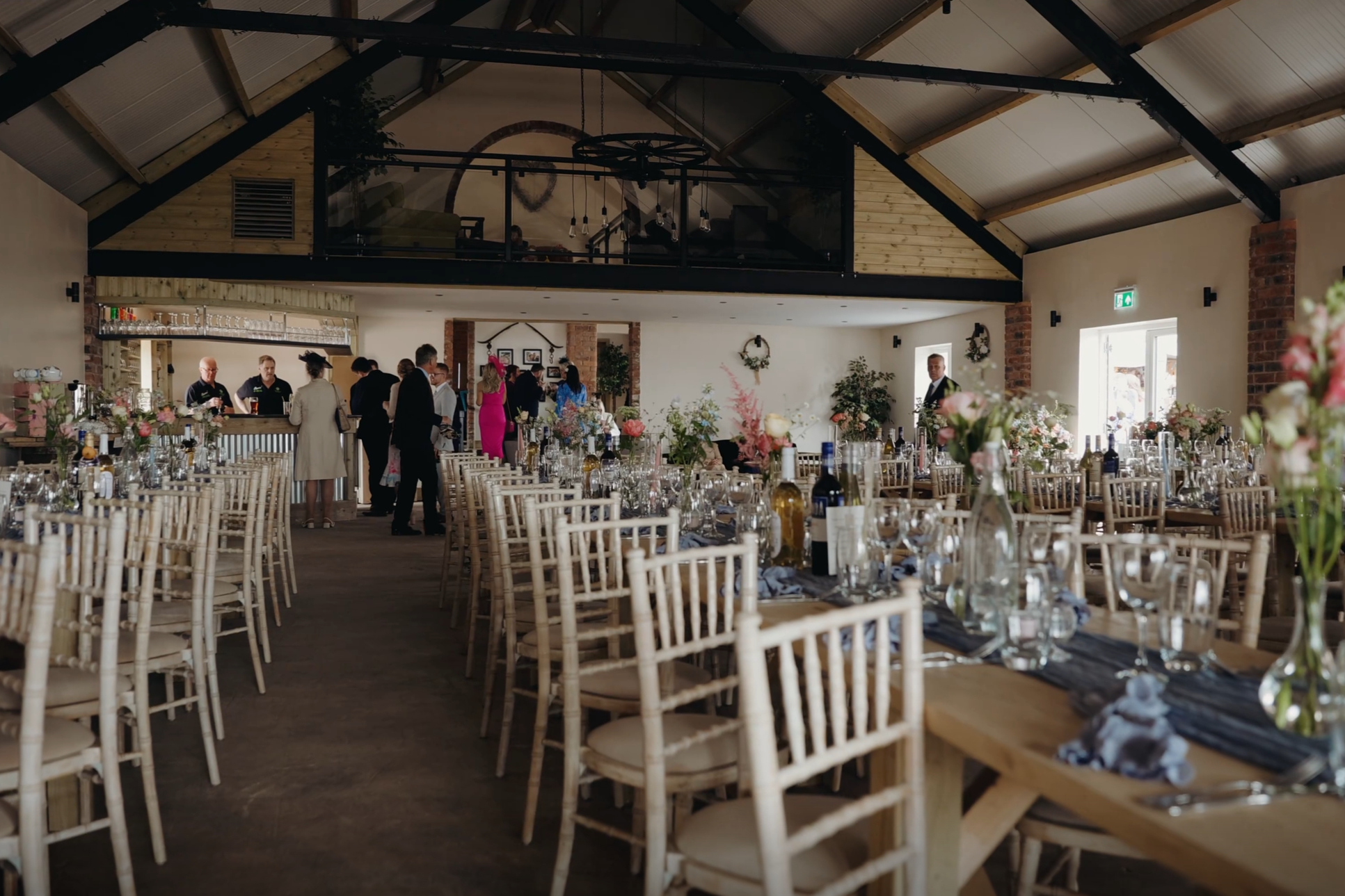 Interior of a decorated event hall with tables set for a celebration, featuring floral centerpieces, wine bottles, and glassware, with people mingling near the bar area and a mezzanine level above.