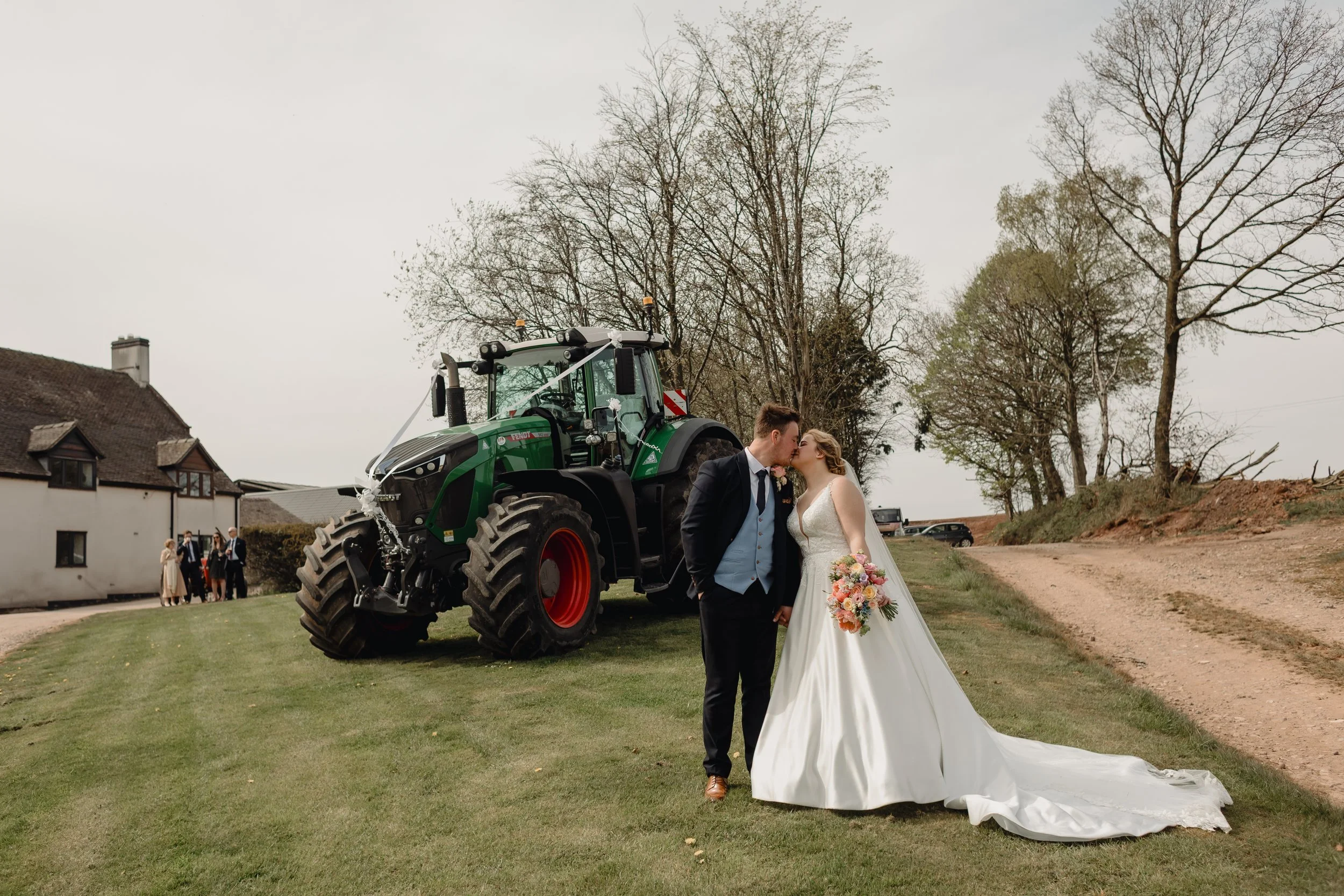 A bride and groom kiss in front of a tractor during their outdoor wedding, with guests and trees in the background.