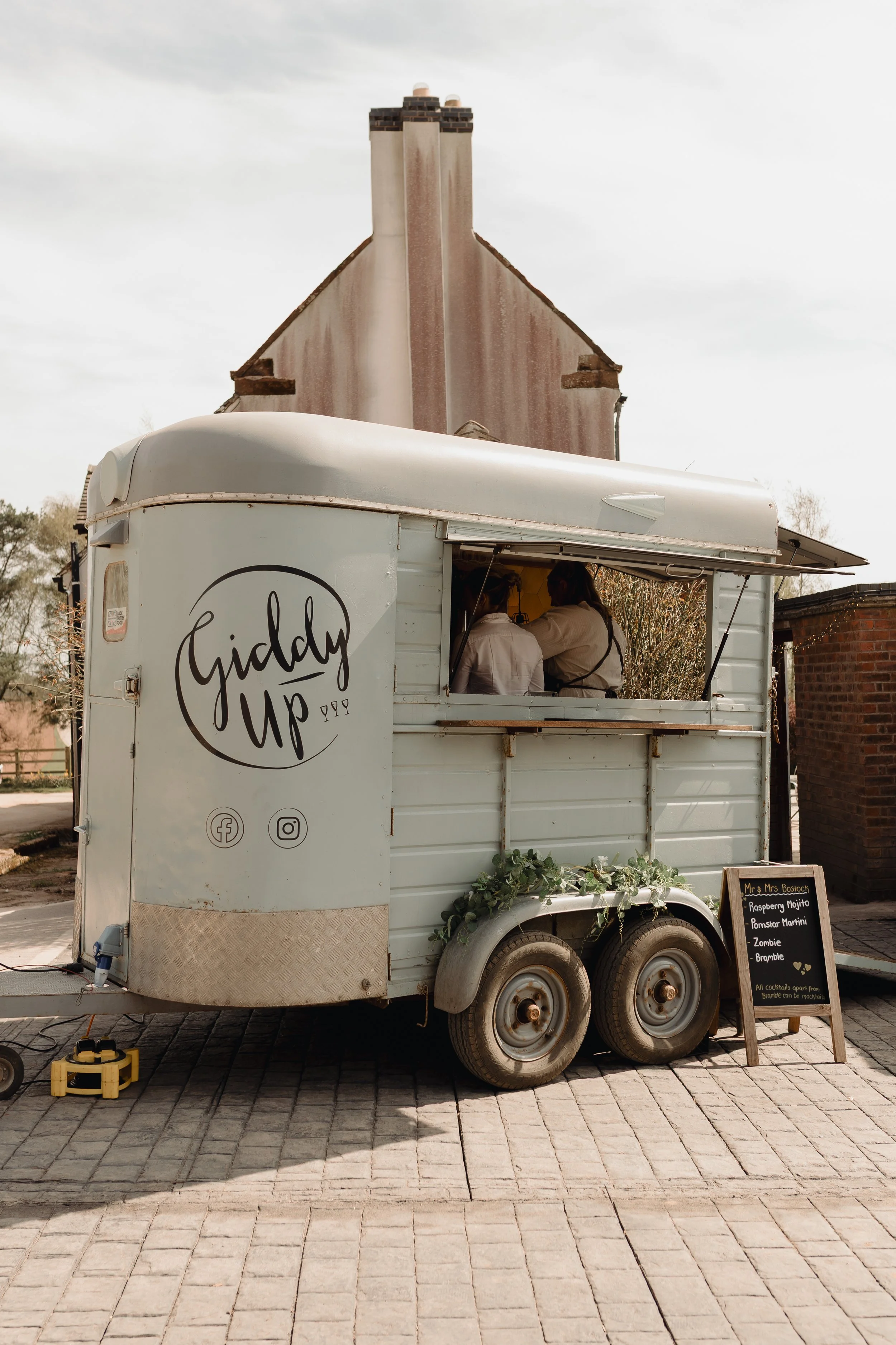 A white food truck named Giddy Up with two people inside, parked on a brick street. A small chalkboard menu is displayed outside, listing drinks such as raspberry mojito, pomegranate martini, zombie, and bramble. The truck is tethered with electrical cords, and there are trees and a brick building in the background.