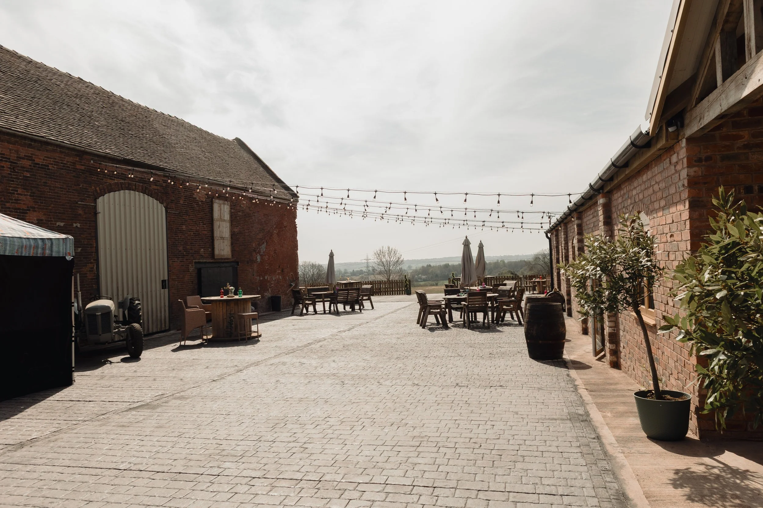 An outdoor patio area between brick buildings with string lights overhead, tables and chairs, umbrellas, potted plants, and a view of the countryside in the distance under a cloudy sky.