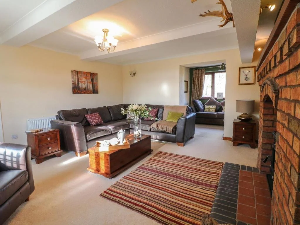 Living room with a large brown leather sofa, a coffee table with a vase of flowers, a striped area rug, a brick fireplace, and additional seating near a window with curtains.
