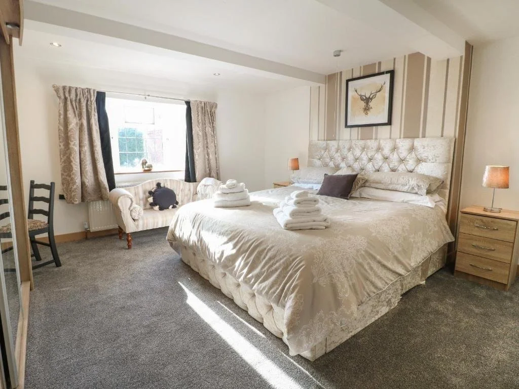 A cozy bedroom with a large bed, beige patterned bedding, and neatly folded towels on top. There are two small wooden nightstands with lamps on each side of the bed. A framed deer head artwork hangs above the headboard. To the left, a window with pat