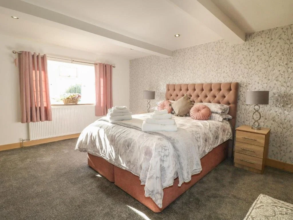 A neatly made bedroom with a large bed with pink tufted headboard, pastel bedding, and decorative pillows. Fluffy towels are folded on the bed. Nightstands with lamps are on each side. Pink curtains frame a window, and patterned wallpaper decorates p