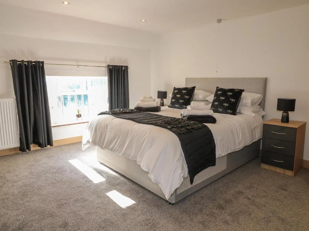 A neatly made bed with white bedding, black and white decorative pillows, and a black throw blanket in a bright bedroom with a window, dark curtains, and two matching black bedside lamps.