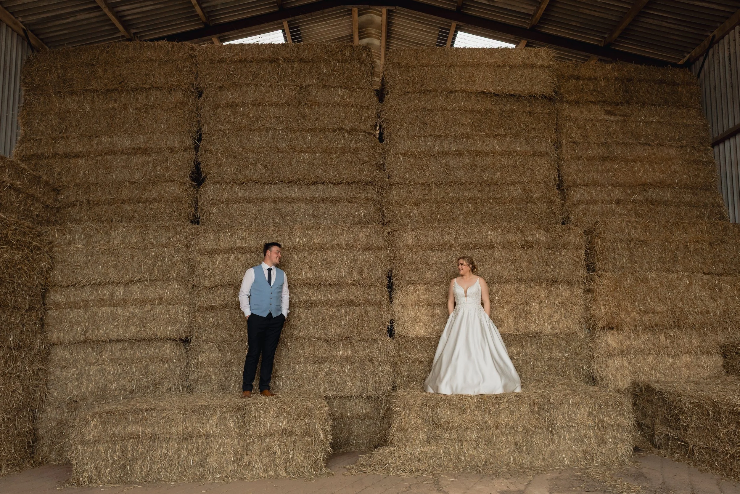 A bride and groom in wedding attire standing in front of a large stack of hay bales inside a barn, with the groom on the left and the bride on the right.