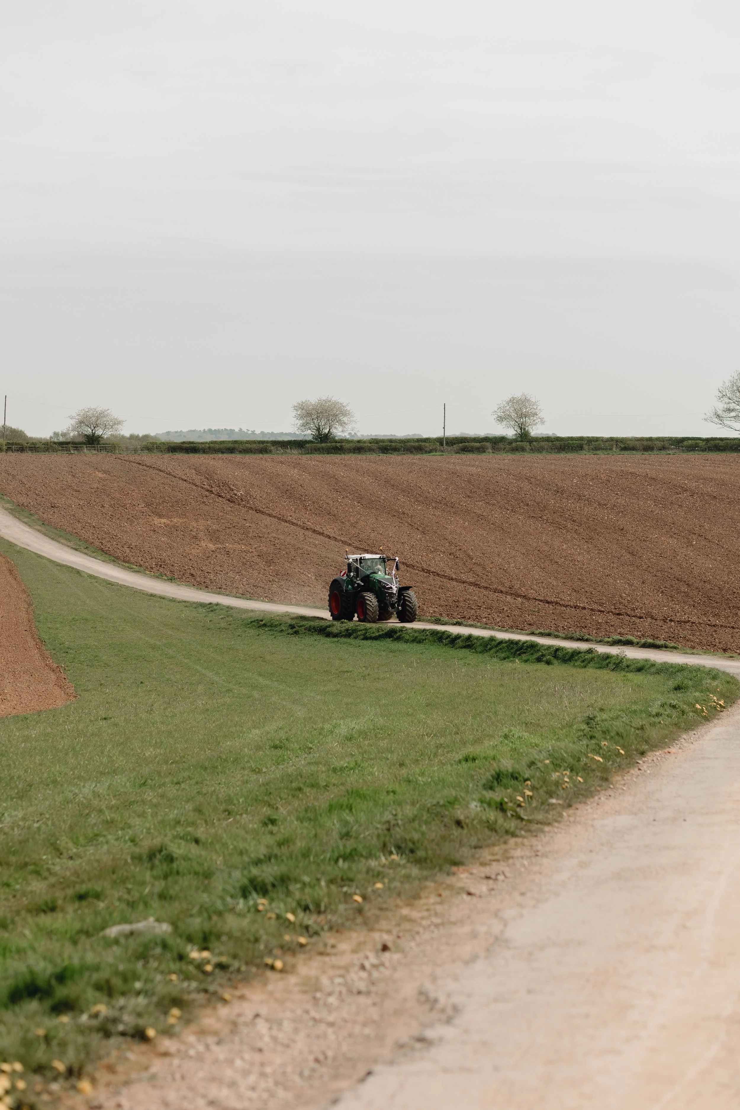 A tractor in a field with tilled soil and a grassy path, trees in the background, under a cloudy sky.
