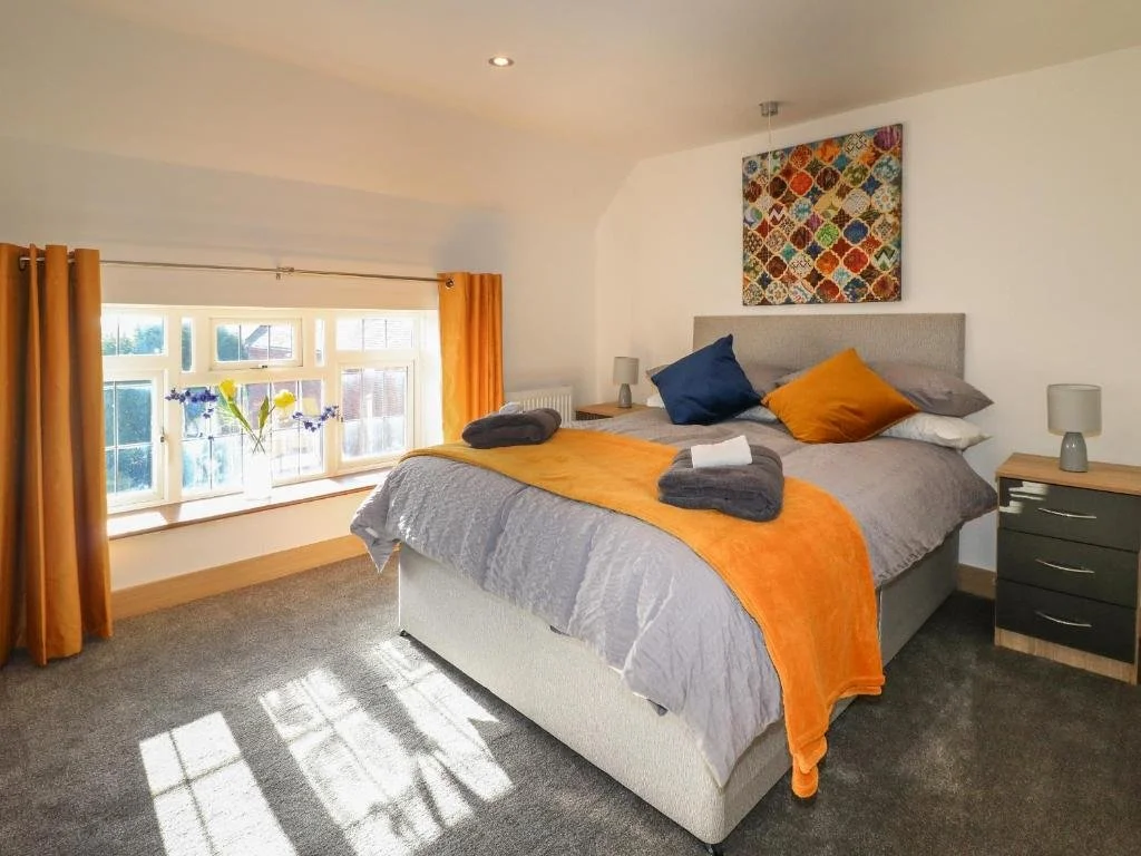 A bedroom with a large bed, orange and gray bedding, and colorful pillows. There are two nightstand lamps, a window with yellow curtains, and a piece of colorful artwork above the bed.