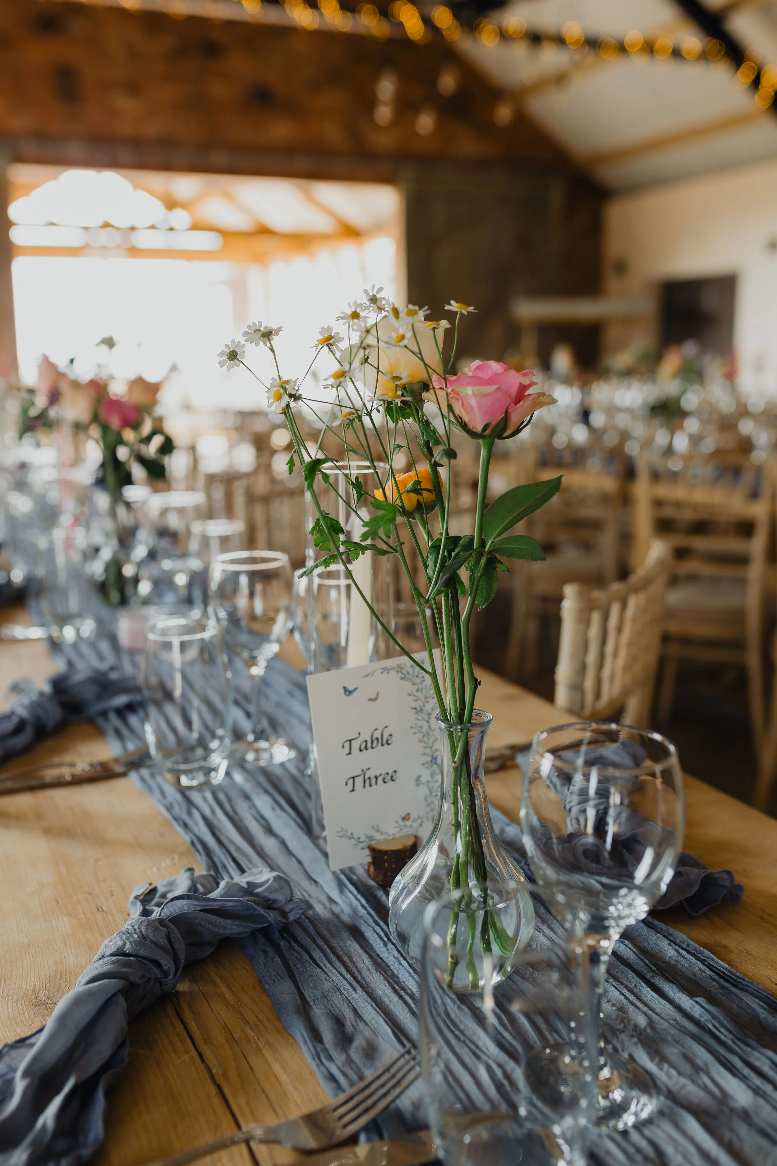 A table setting with a small vase holding pink and white flowers, surrounded by wine glasses, utensils, and a gray table runner in a rustic indoor venue with wooden chairs and warm lighting.
