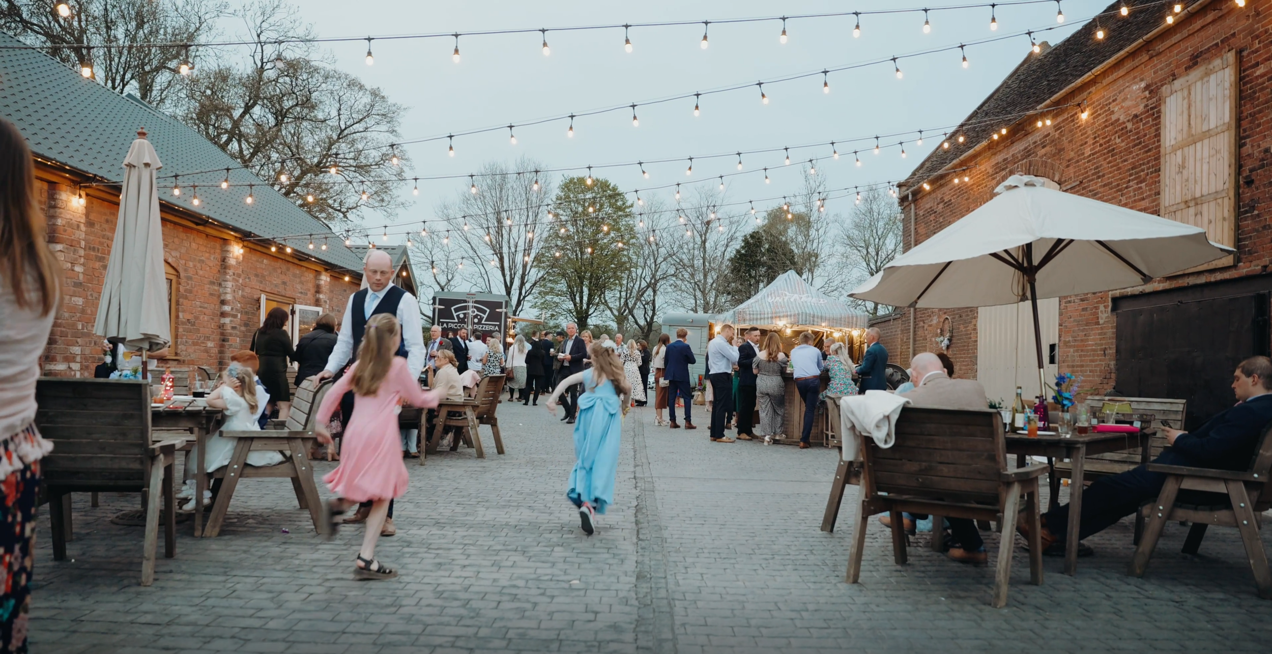 People are gathering and socializing outdoors at a festive event in a courtyard with string lights, brick buildings, and trees in the background.