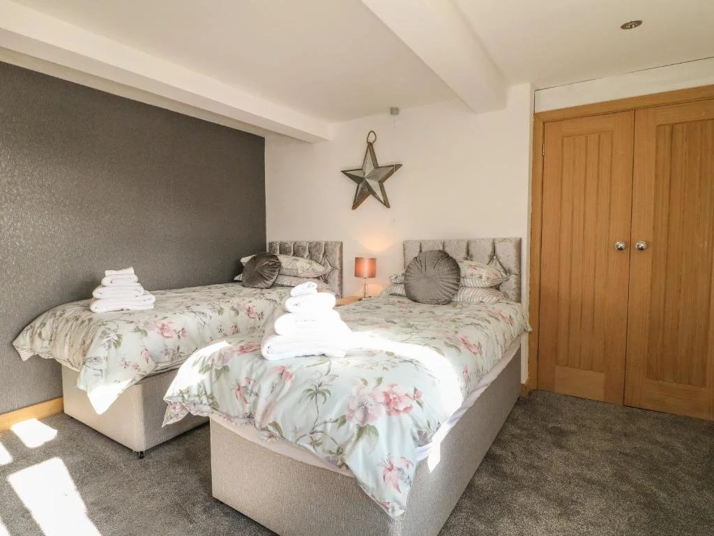 A bedroom with two beds with floral bedding, grey headboards, and two stacks of towels on each bed. There is a star decoration on the wall, a small bedside lamp, and a wooden closet door.
