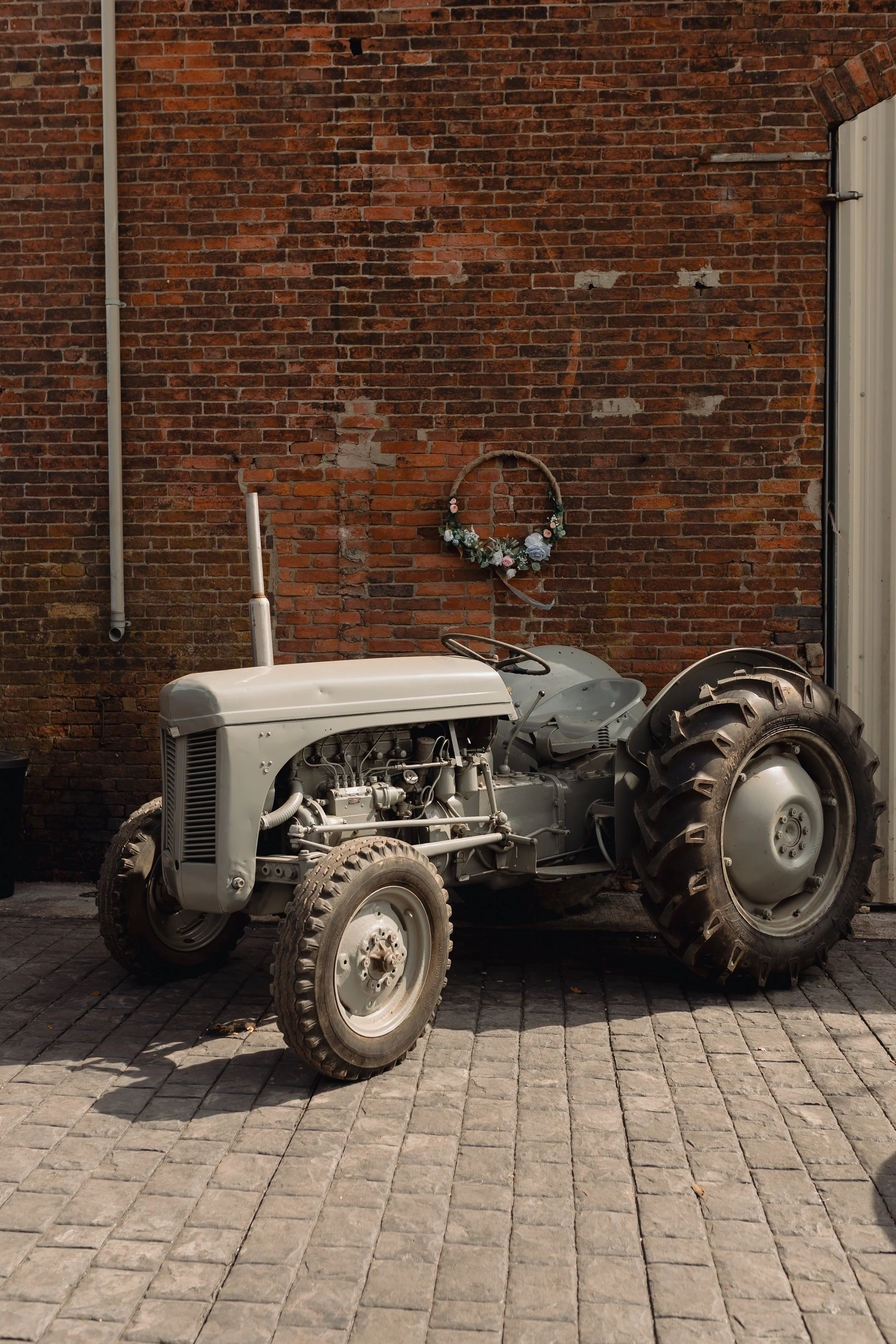 A partially assembled vintage tractor in front of a brick wall with a floral wreath hanging on the wall and a pipe running vertically.