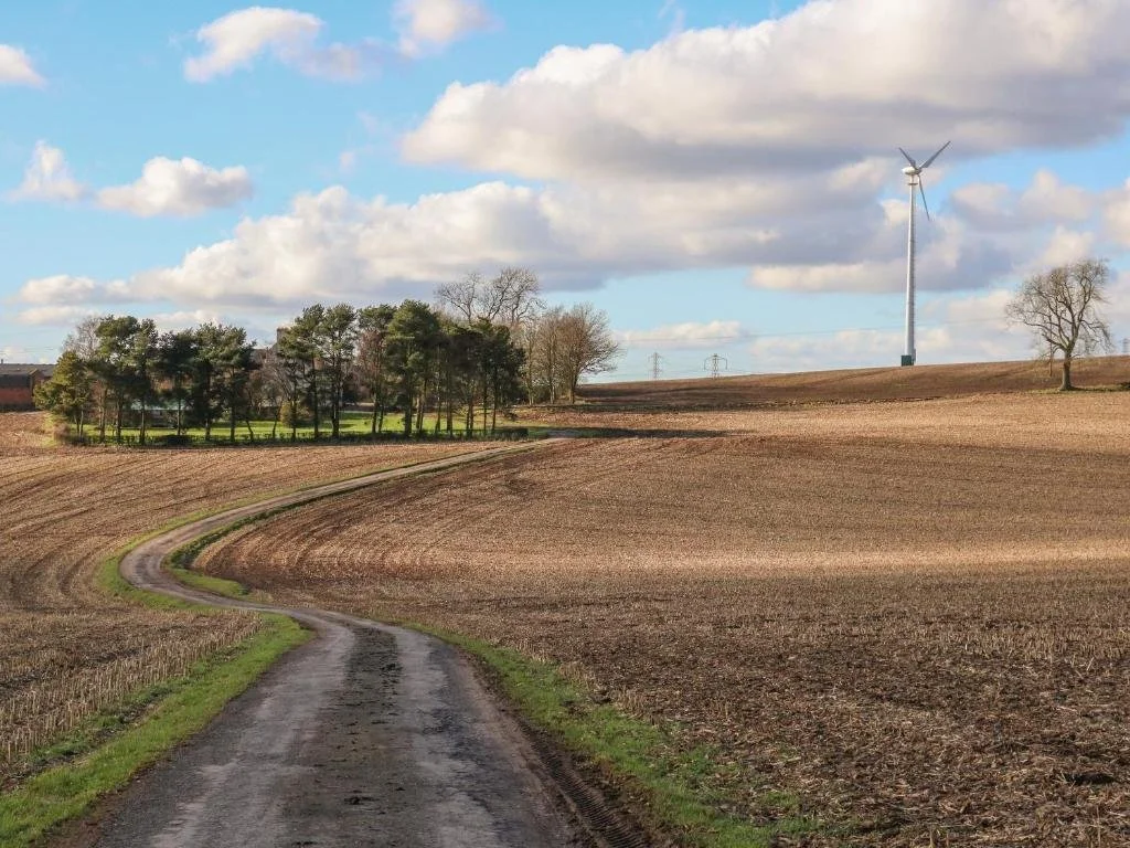 A rural landscape with a winding dirt road, plowed fields on either side, a small cluster of trees and a house, and a large wind turbine in the distance under a partly cloudy sky.