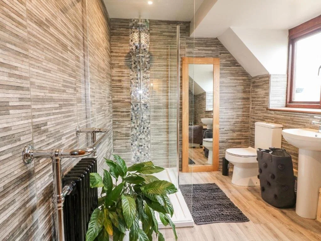 A modern bathroom with wooden and tiled walls, including a walk-in shower with mosaic tiles and glass door, a small mirror framed in wood, a toilet, a pedestal sink, a window, and a plant on a radiator.