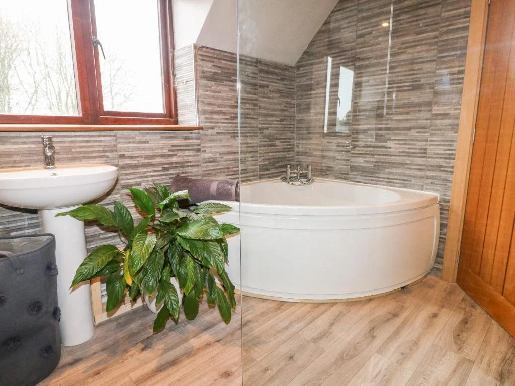Bathroom with a corner bathtub, a pedestal sink, a window, a potted plant, and wooden and tiled wall and flooring.