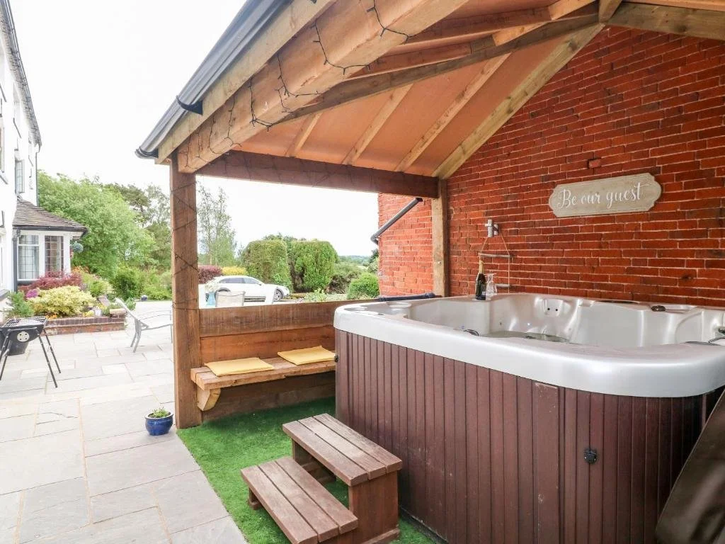Outdoor hot tub under a wooden roof with a brick wall, a sign saying 'Be our guest,' and a view of a driveway with cars and green trees.