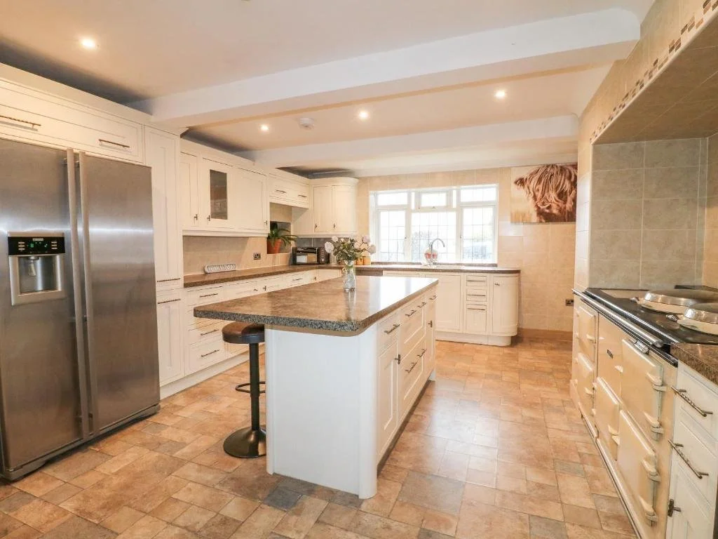 A spacious kitchen with white cabinets, a large island with a granite countertop, a stainless steel refrigerator, and a window above the sink, with tiled flooring and recessed lighting.