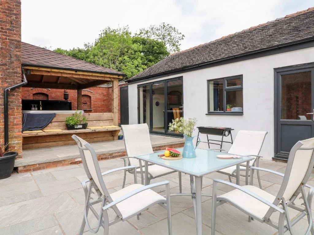 Outdoor patio with a glass table surrounded by five white chairs, a vase with flowers, and a plate of fruit. Behind is a white building with black window and door frames, and a small barbecue grill. There is also a covered brick area with a built-in 