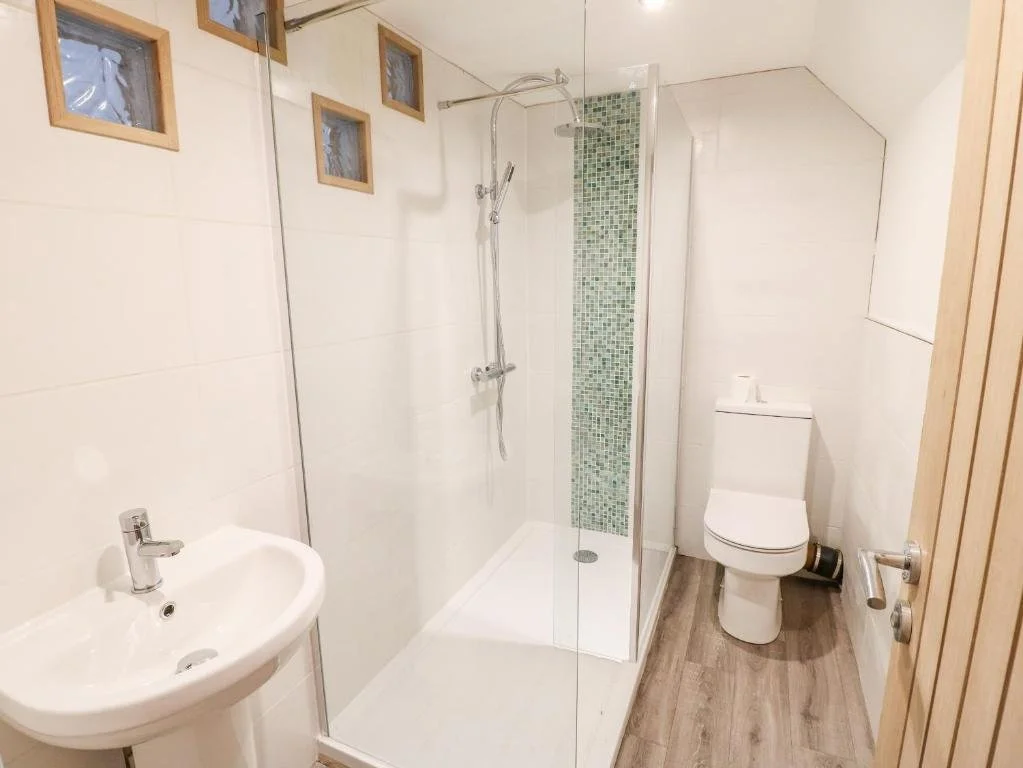 Small bathroom featuring a corner shower with glass panels, white sink, and white toilet, with wood accents and small decorative windows.
