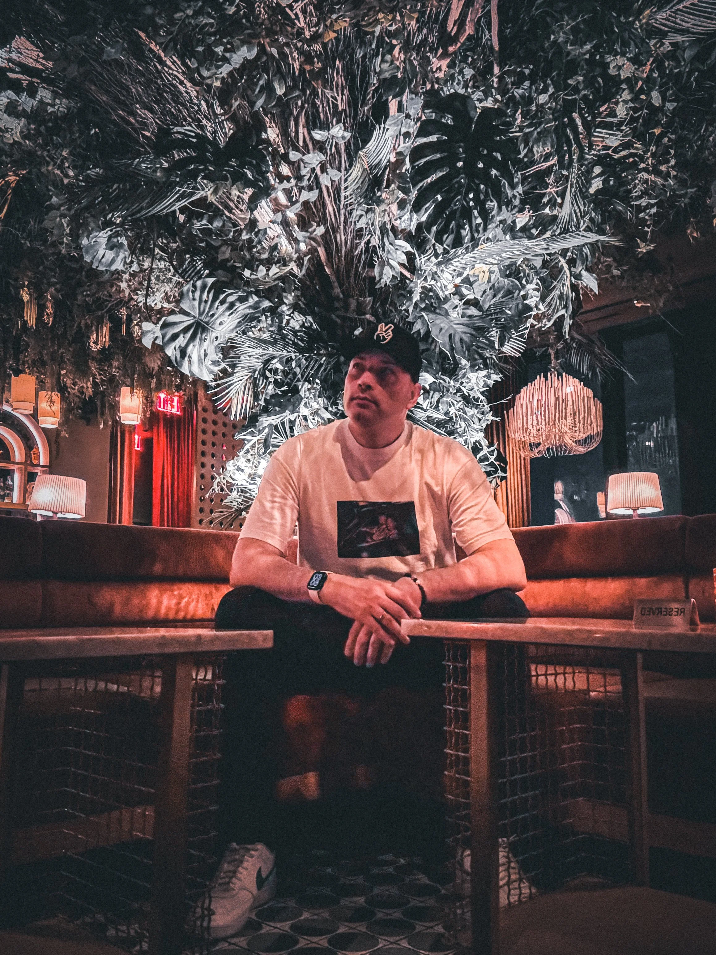 The Professor sitting on a brown leathery sofa in a dimly lit, modern interior with botanical decor overhead and ambient lighting.
