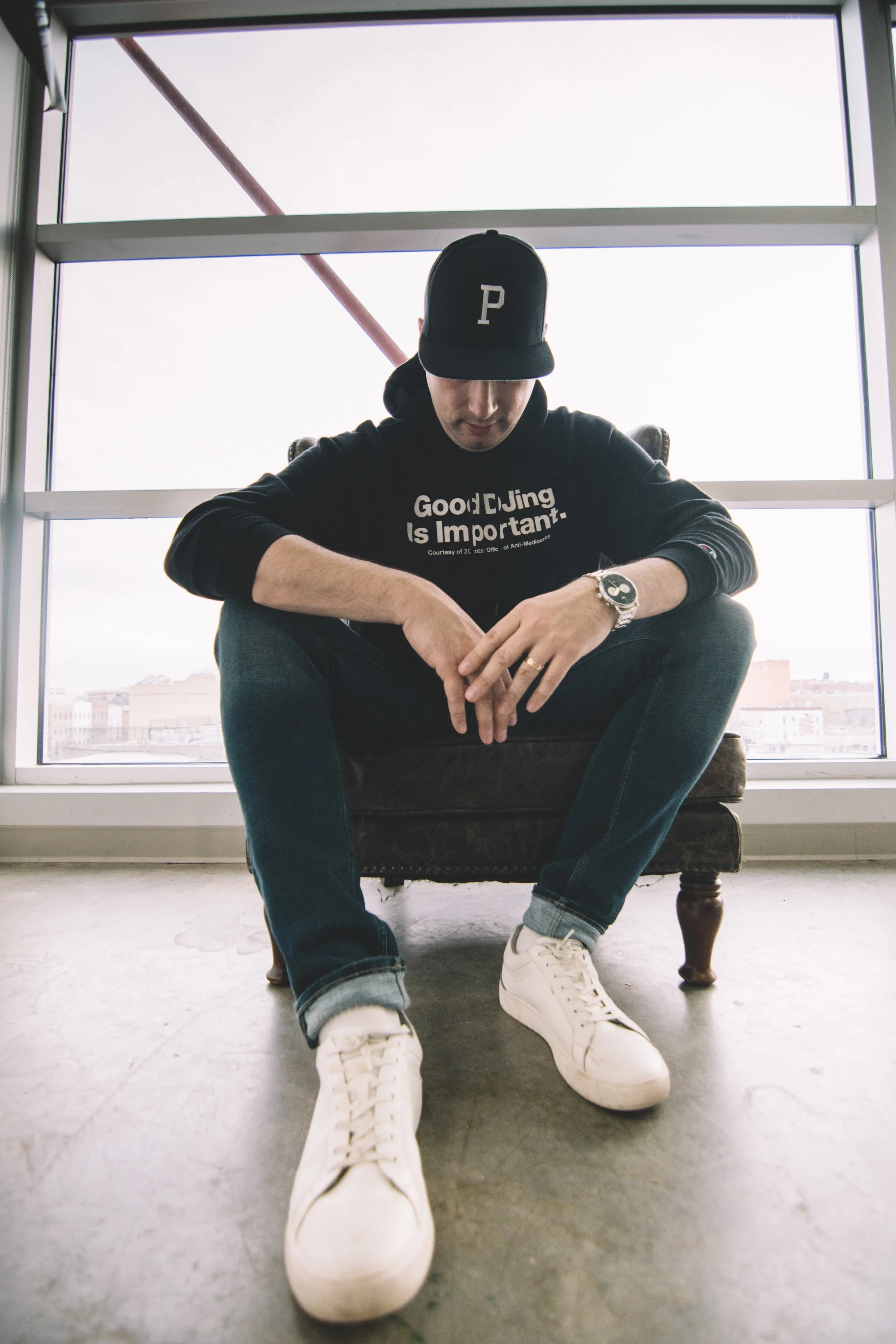 The Professor sitting on a vintage armchair, wearing a black hoodie with white text, a black baseball cap with a 'P' logo, white sneakers, and a wristwatch, positioned in front of a large window with a cityscape view.