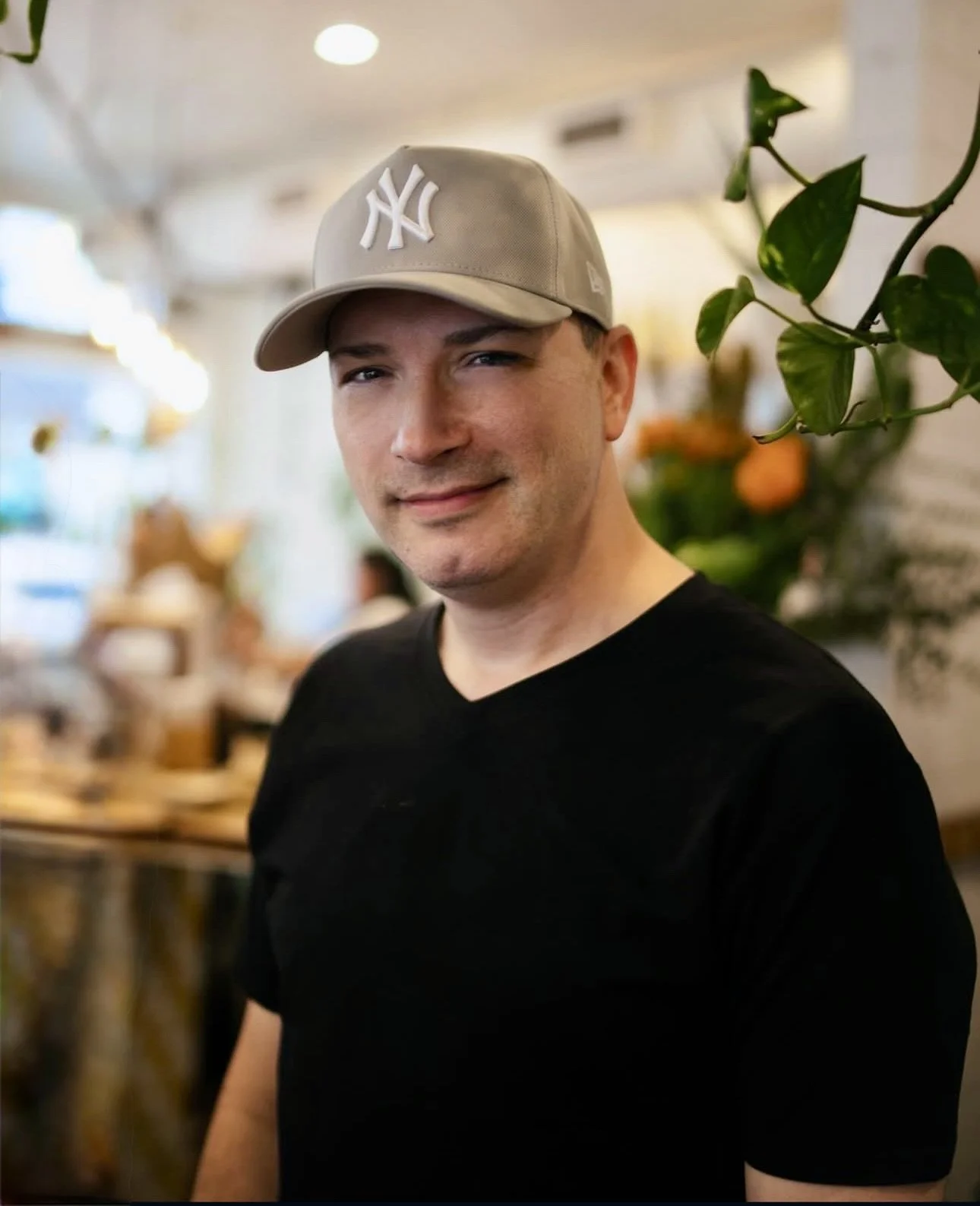 The Professor wearing a beige New York Yankees baseball cap and a black t-shirt, smiling in a restaurant or cafe with blurred background and green plants.