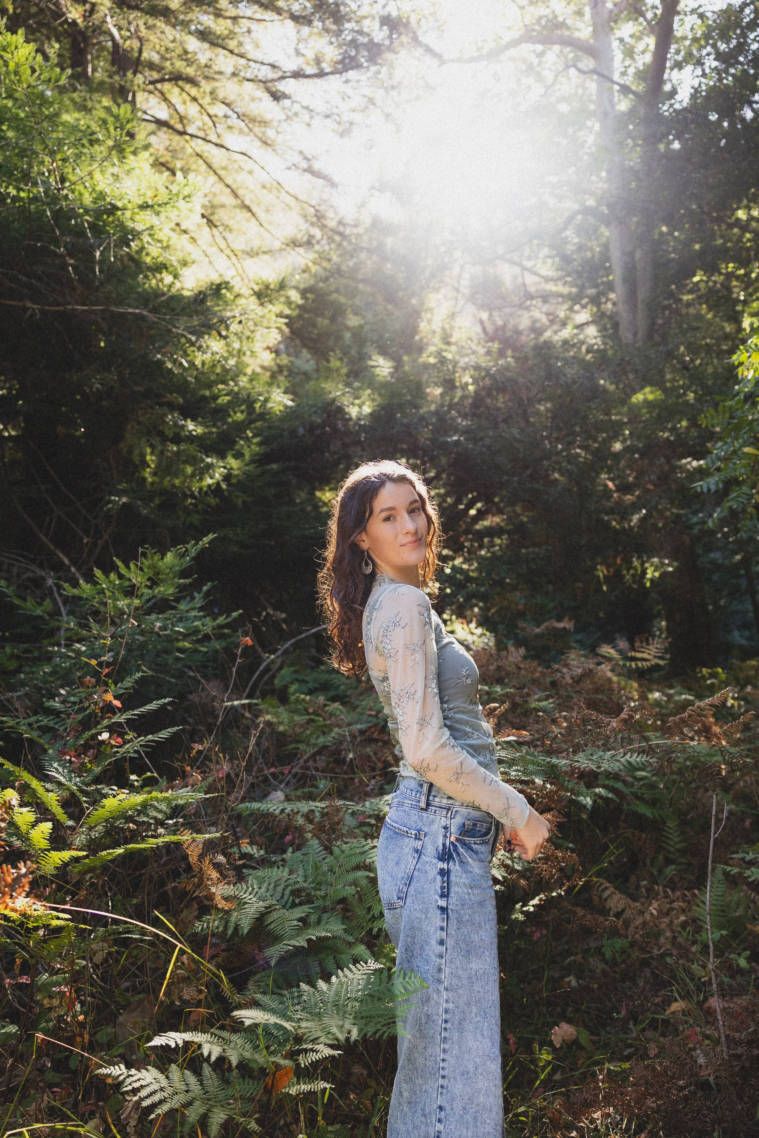 A young woman with wavy brown hair standing in a forest with sunlight filtering through trees, wearing a light-colored long-sleeve top with floral designs and high-waisted jeans.