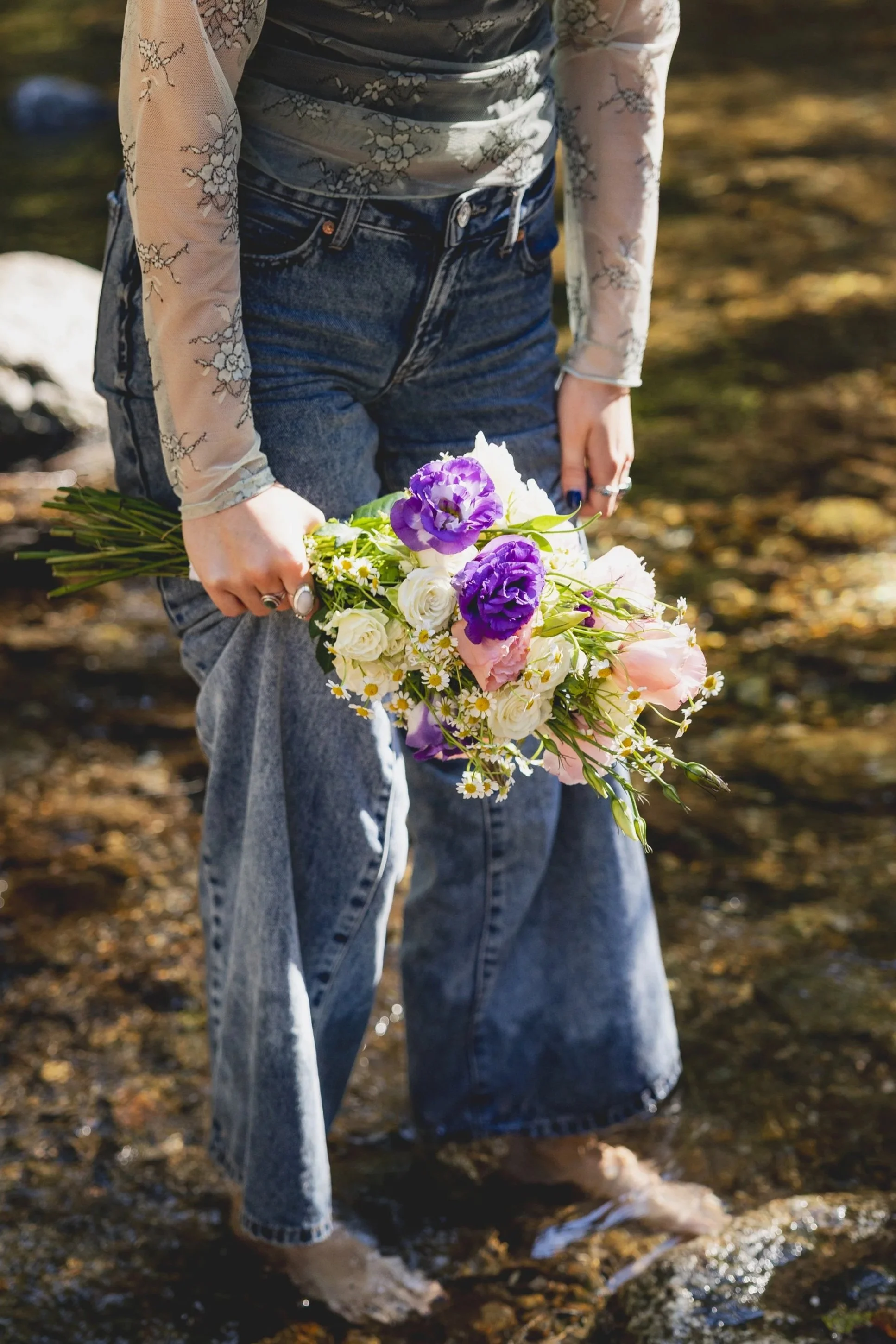 Person wearing jeans and a sheer, embroidered top holding a colorful bouquet of purple, white, and pink flowers while standing barefoot in a shallow stream.