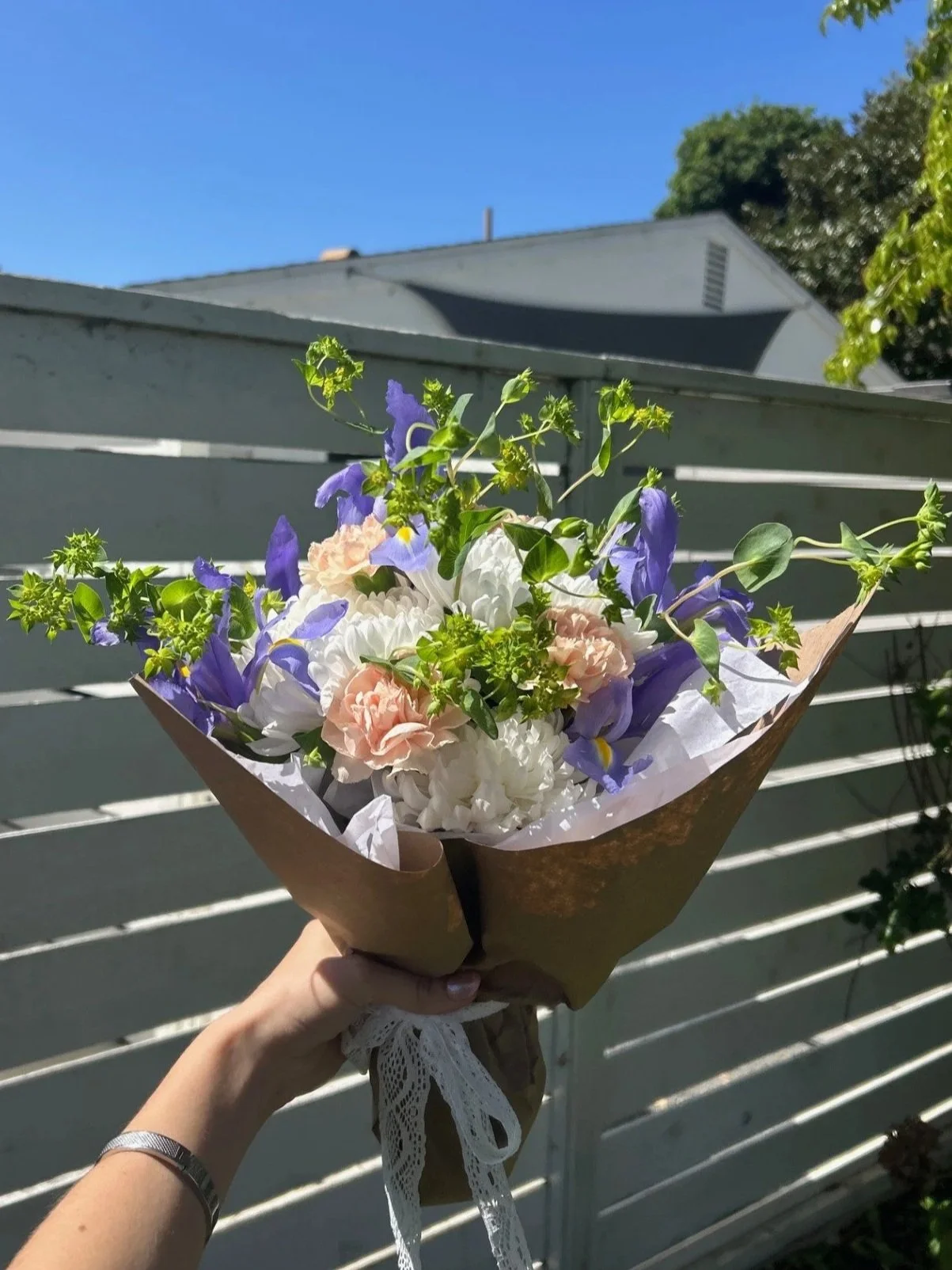 A hand holding a bouquet of white, pink, and purple flowers against a blue sky and a white fence.