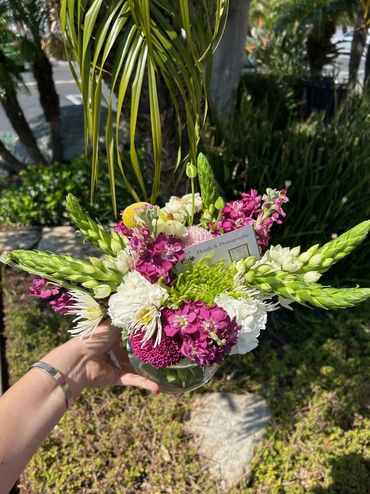 A hand holding a colorful floral bouquet outdoors, featuring white, pink, and green flowers with some greenery, sunlight, and a garden background.