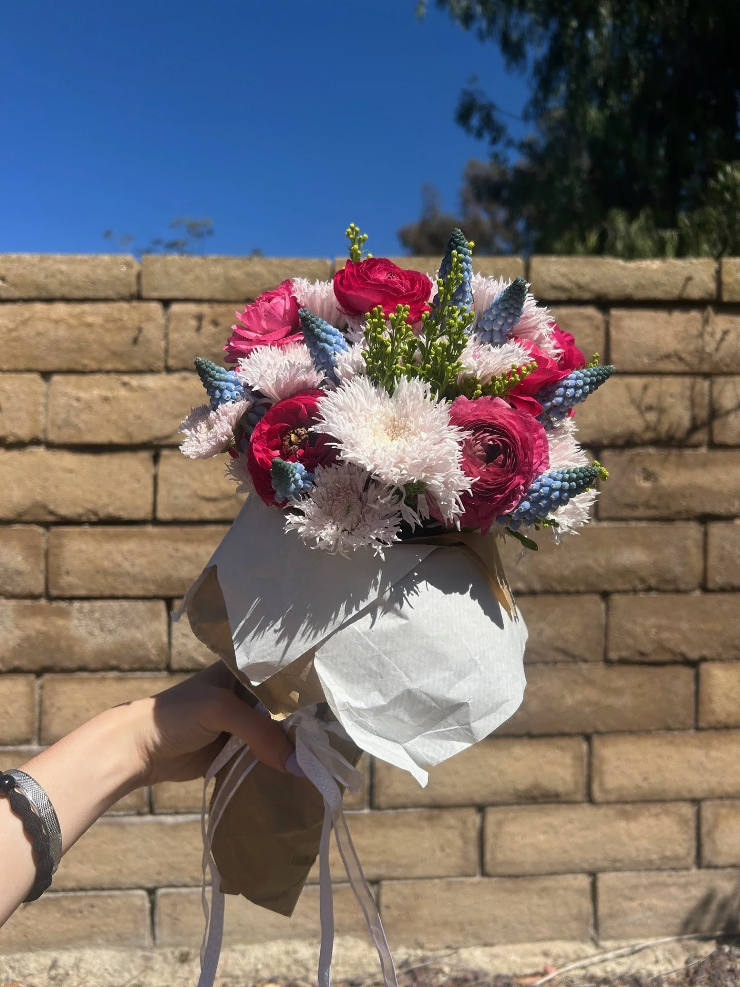 A bouquet of pink, white, and red flowers with blue accent flowers, held against a brick wall with a clear blue sky in the background.