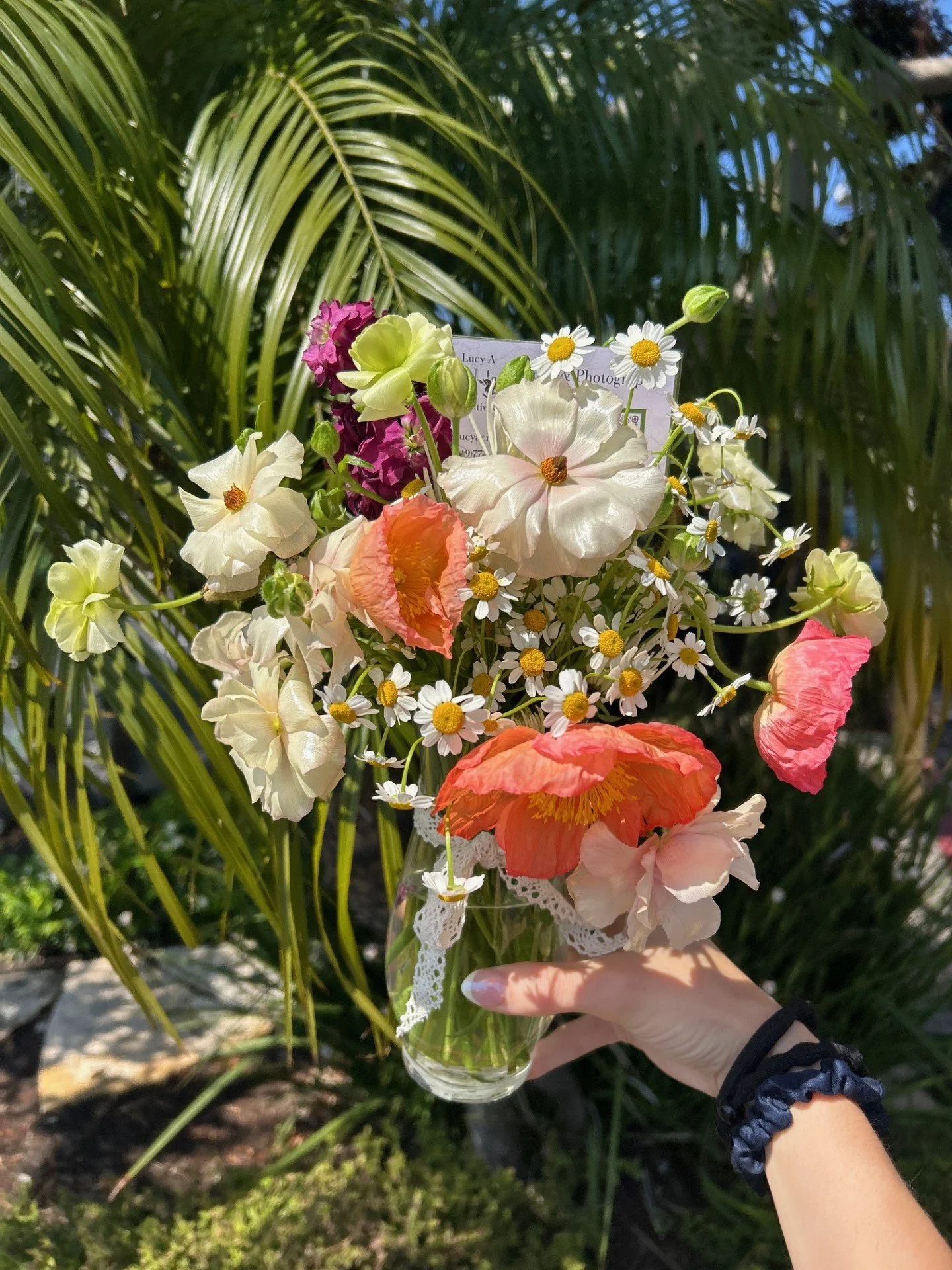 A hand holding a glass vase with a bouquet of various colorful flowers against a background of green palm leaves.