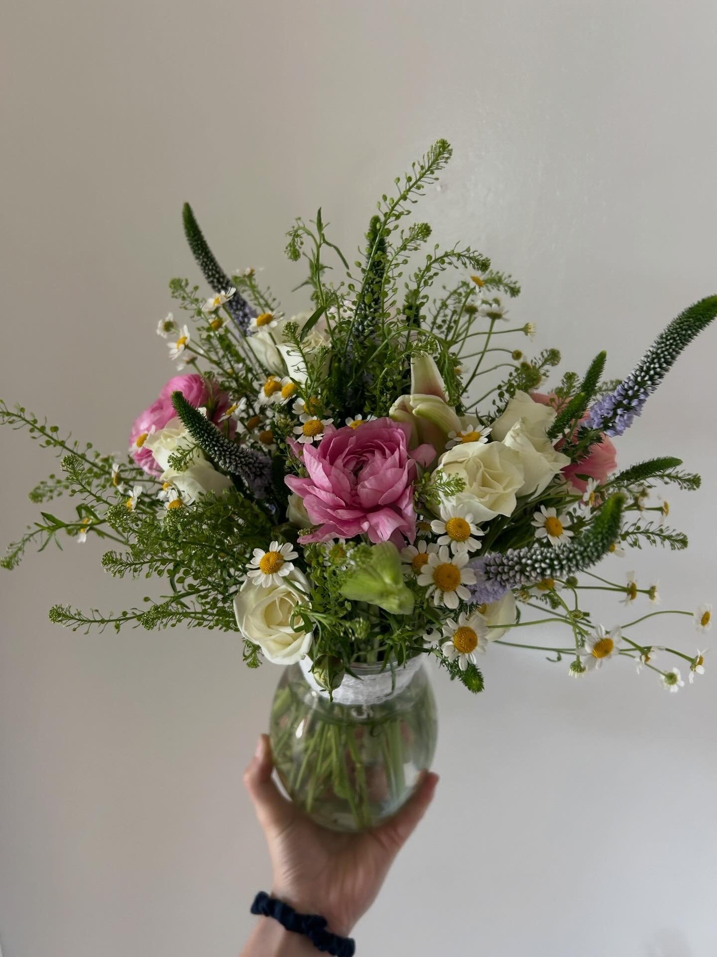 Hand holding a glass vase with a mixed bouquet of pink and white flowers, daisies, and green foliage against a plain background.