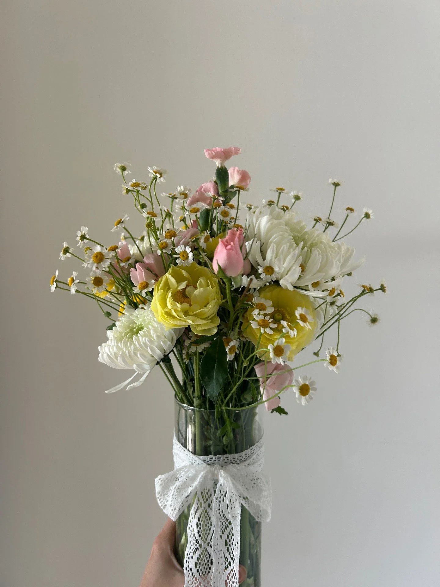 A hand holding a glass vase with a bouquet of various flowers, including white and yellow chrysanthemums, pink tulips, and small white daisies with yellow centers, decorated with a white lace ribbon.