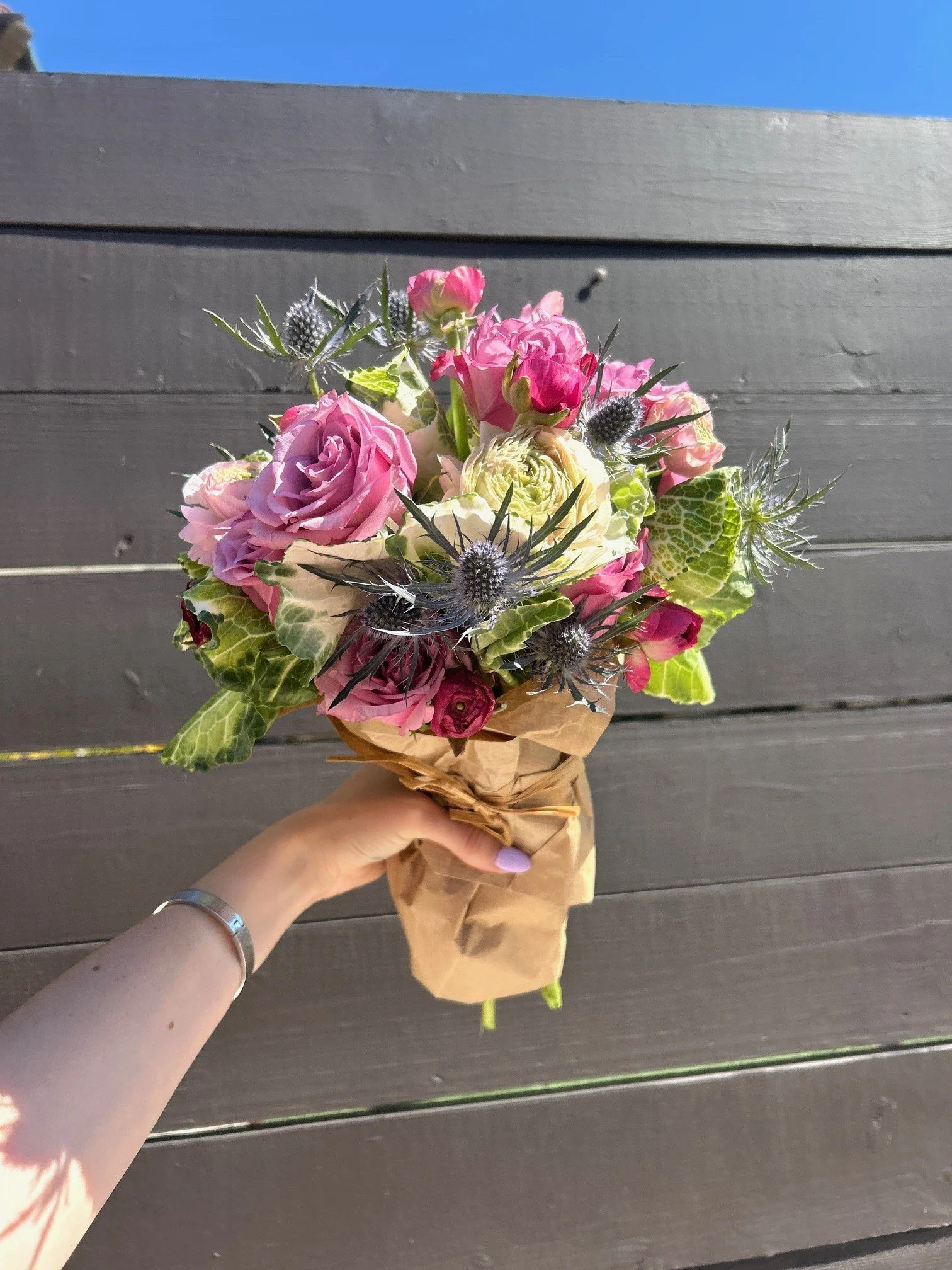 Hand holding a bouquet of pink and white flowers wrapped in brown paper, against a dark wooden fence and blue sky background.
