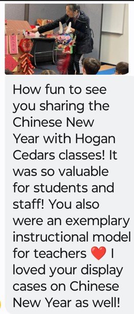 A teacher and a student share a moment during a Chinese New Year celebration in a classroom. The teacher is holding a colorful display, and there are festive decorations on the table. Several students are watching.