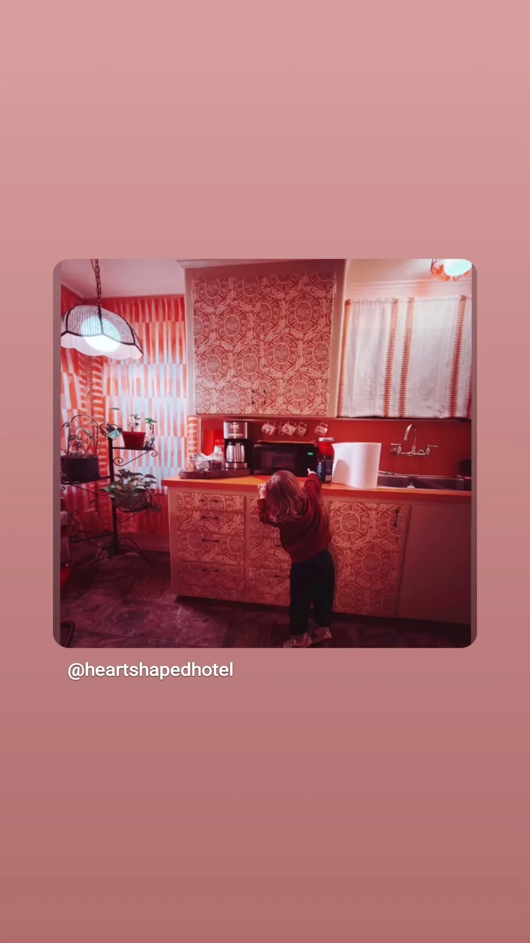 A young girl reaching for items on a kitchen counter in a warm red-toned kitchen with patterned wallpaper, curtains, and appliances.