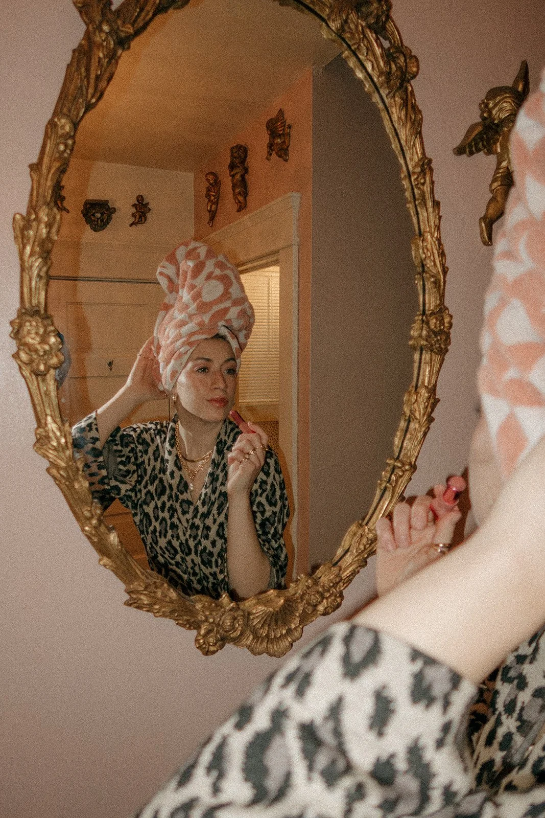 A woman looking into an ornate gold mirror, wearing a leopard print robe and pink towel turban, applying lipstick.