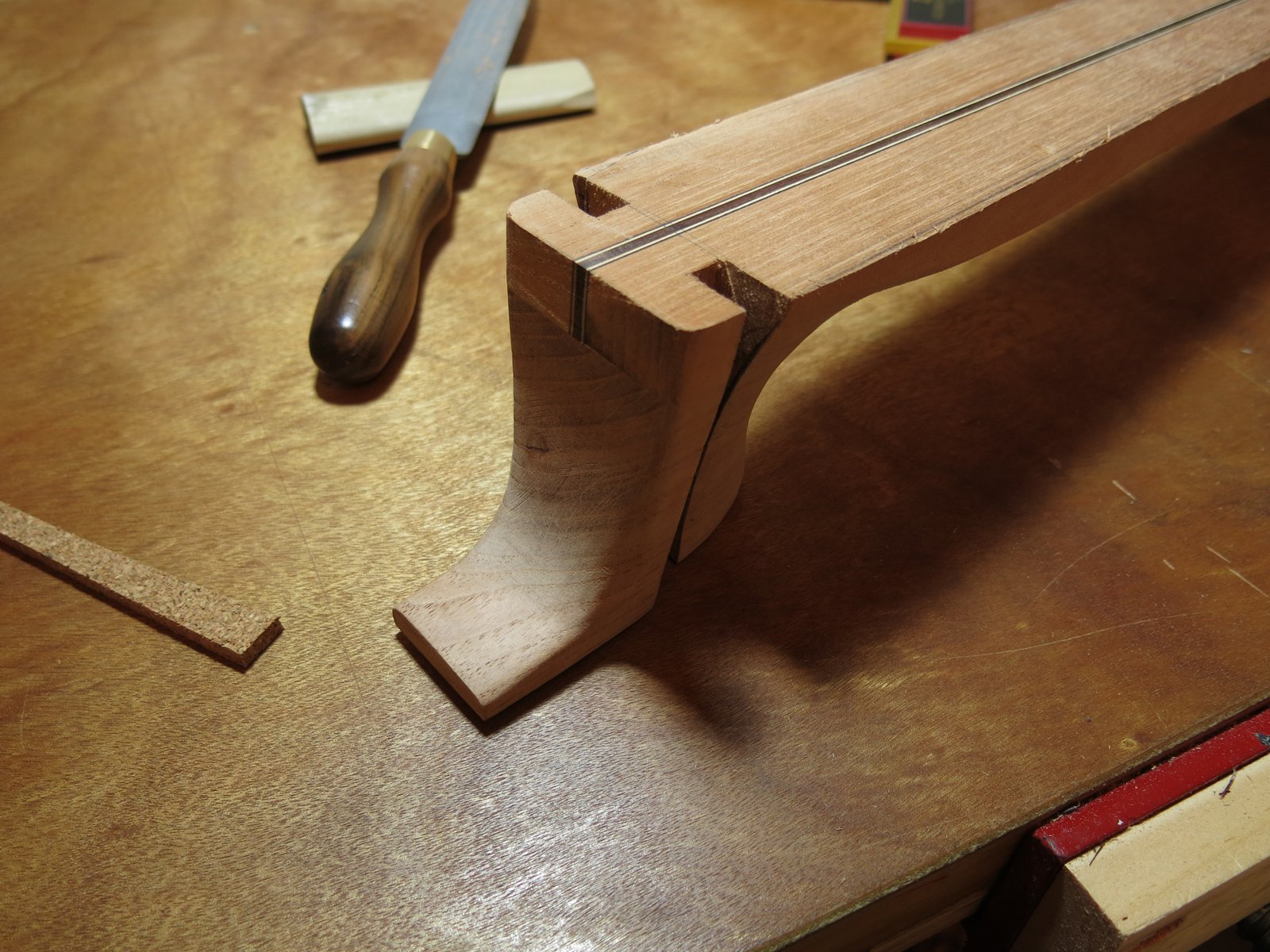 A woodworking project in progress with a partially assembled wooden guitar neck, a chisel, and a piece of cork on a wooden workbench.