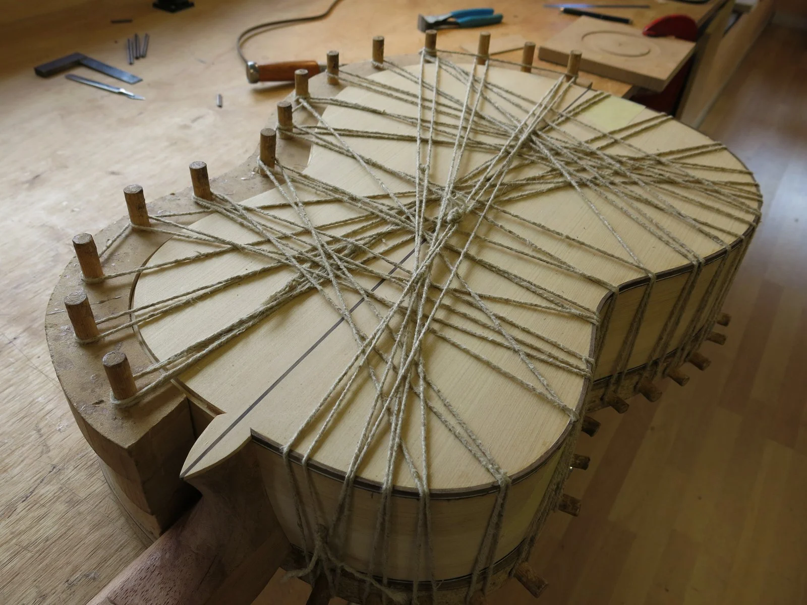 A wooden guitar body being assembled with numerous strings stretched across, secured by wooden pegs, on a workbench in a woodworking shop.
