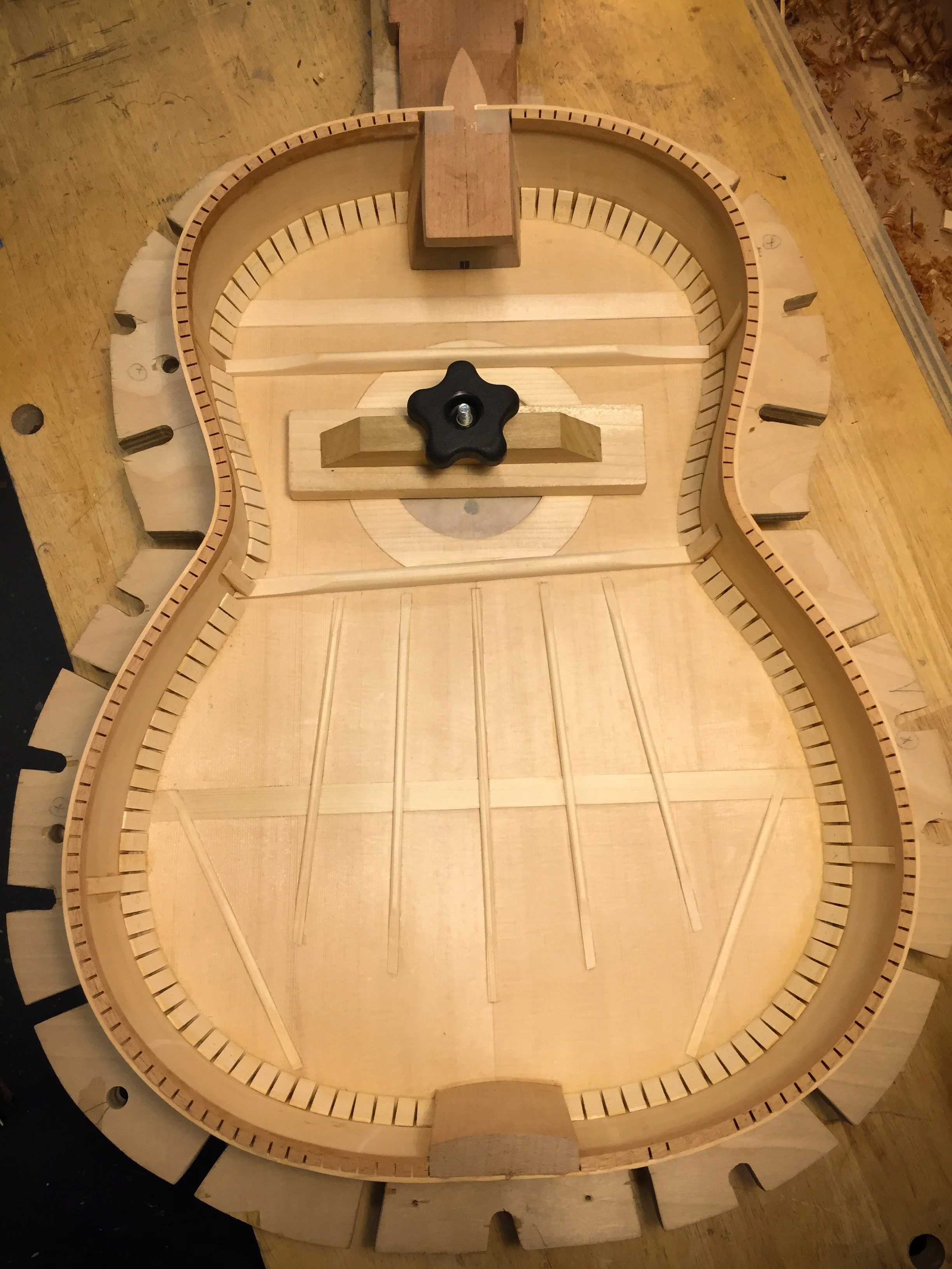 Empty wooden guitar body with internal bracing, a central tuning mechanism, and a black plastic knob on a workbench.