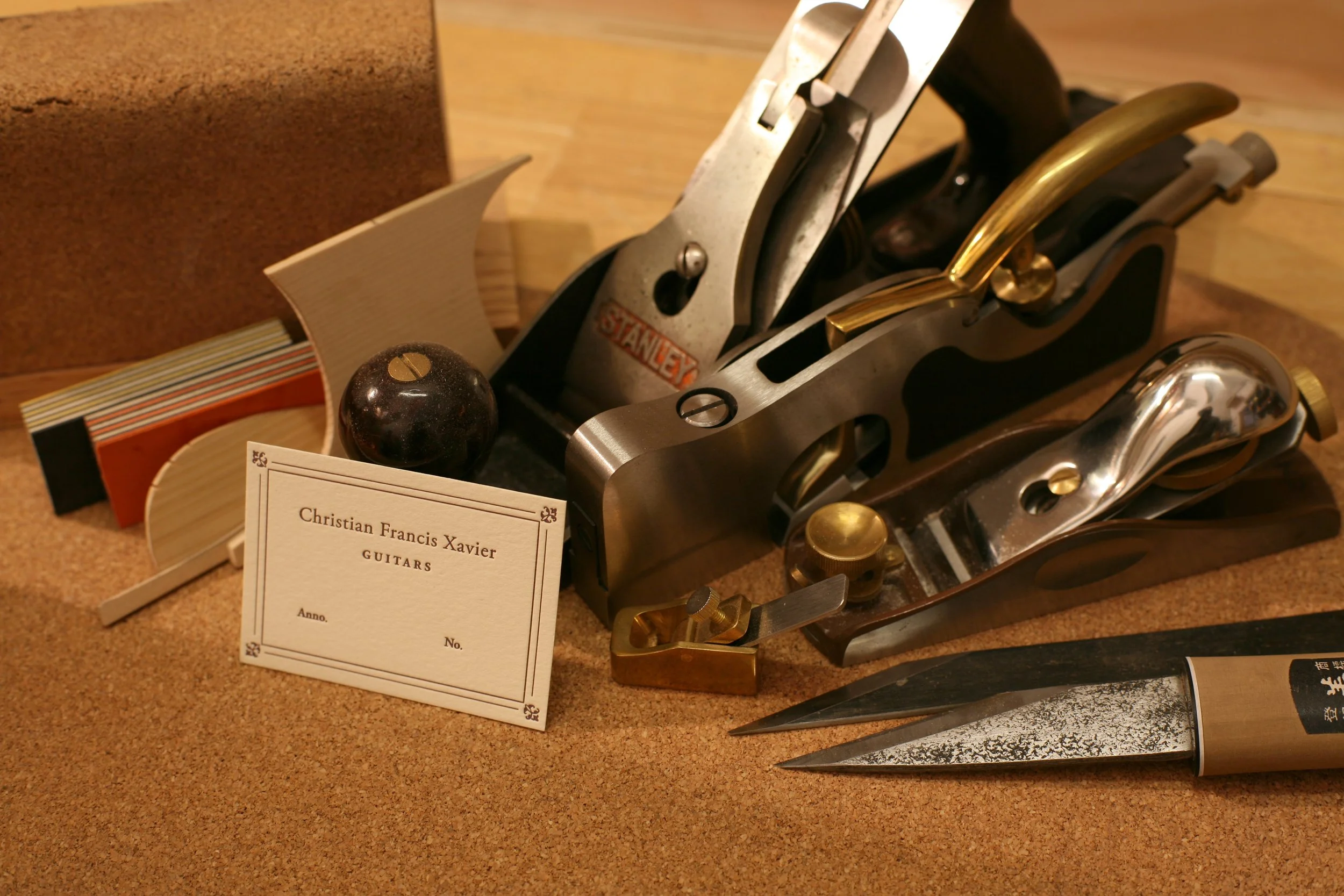 A collection of woodworking tools including a hand plane, carving knives, a sanding block, and a card for Christian Francis Xavier Guitars, arranged on a wooden surface.