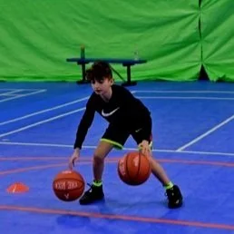Young boy practicing basketball drills on an indoor court with a green screen background, dribbling two basketballs simultaneously.