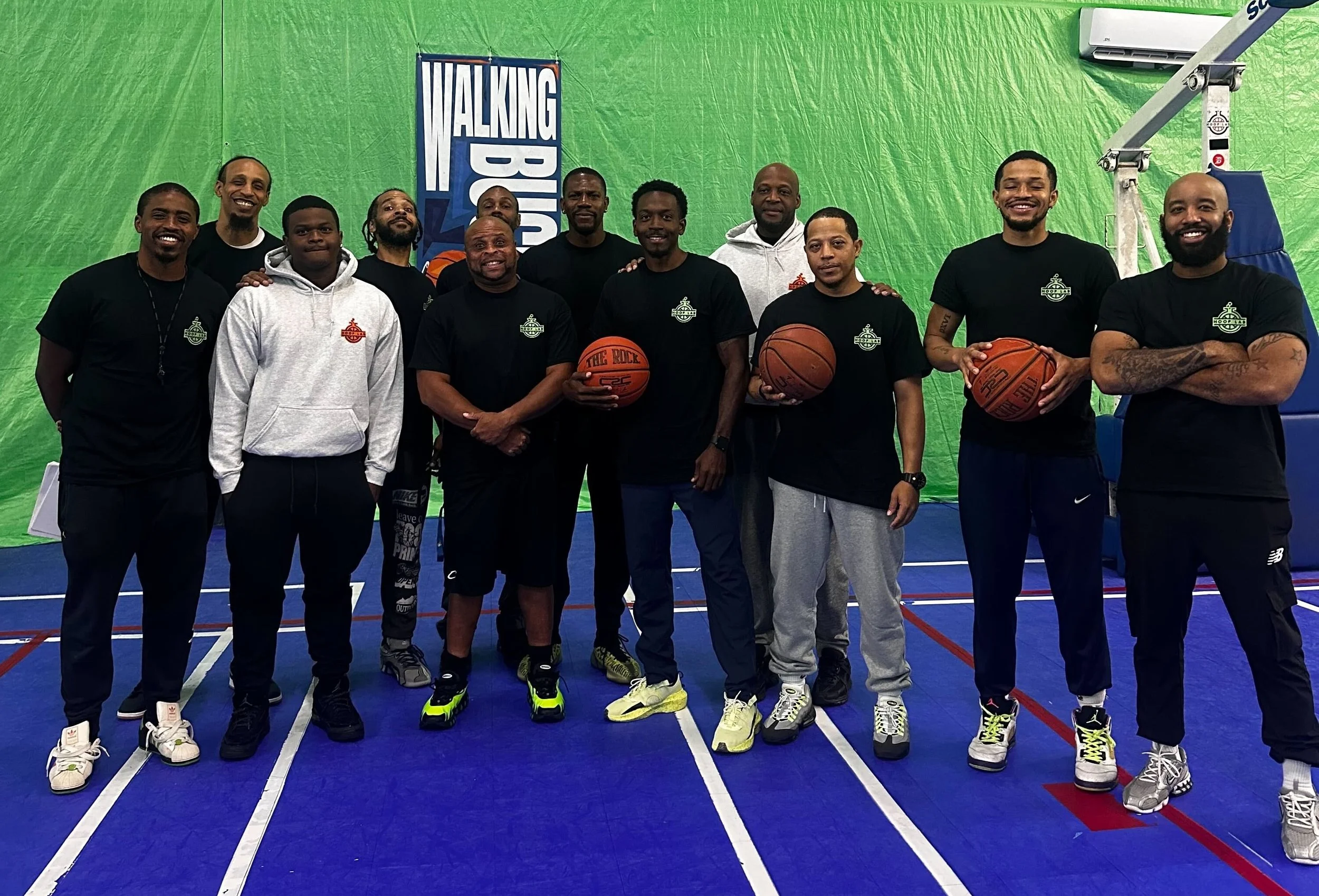 Group of men standing on a basketball court, some holding basketballs, with a green backdrop and a Hoop Lab banner