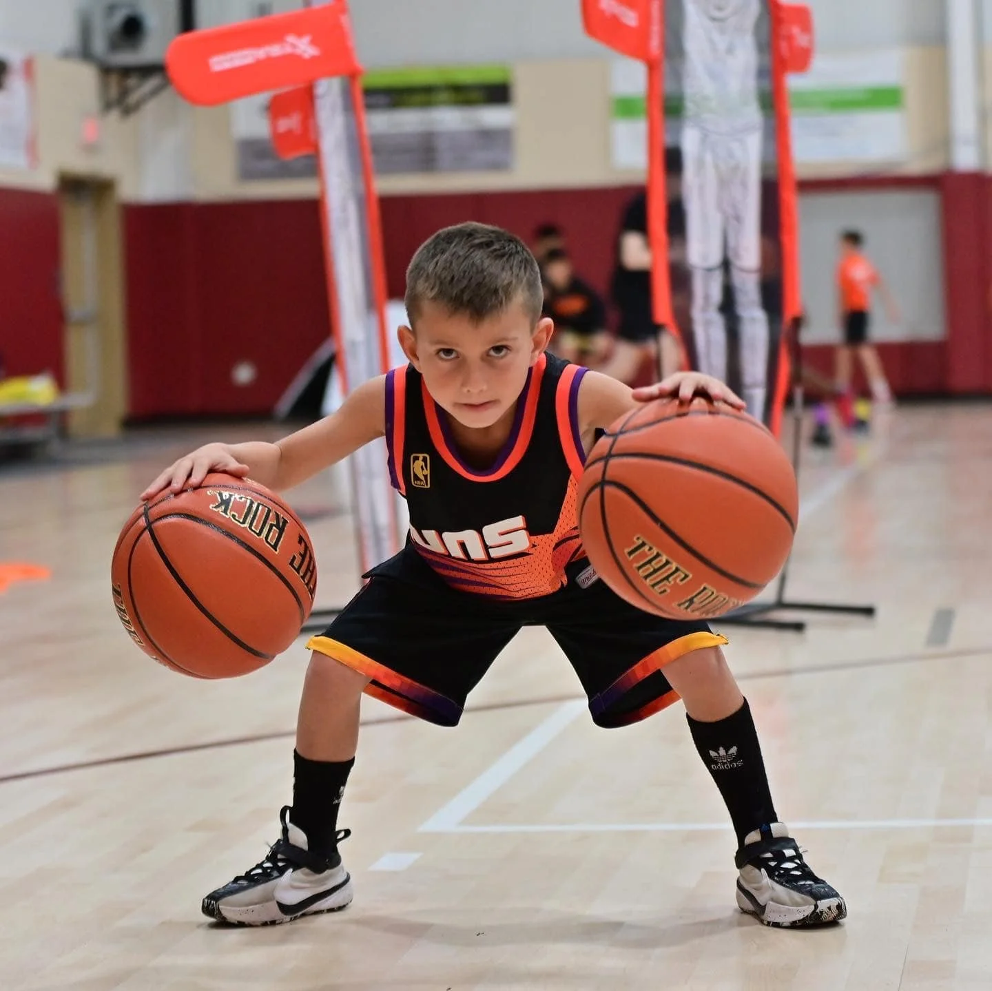 Young boy playing basketball in an indoor gym, holding a basketball in each hand with a focused expression.