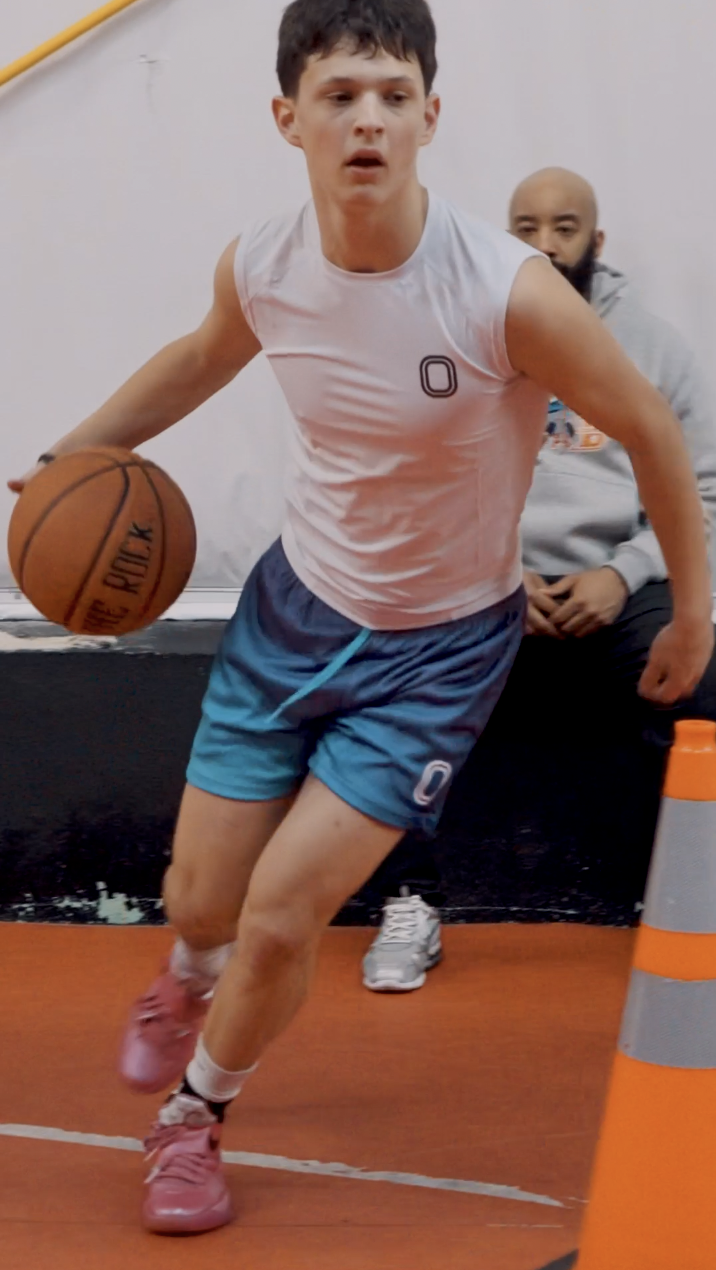 Young male basketball player dribbling a basketball indoors, wearing a white tank top and blue shorts with a number 0, with another person sitting on the bench in the background.